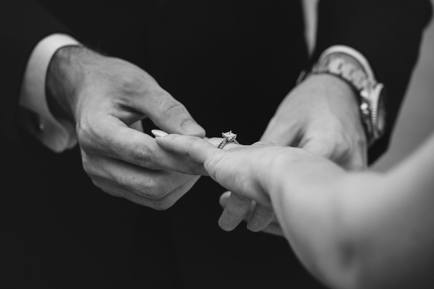 Close-up black and white photo of groom placing diamond engagement ring on bride's finger.