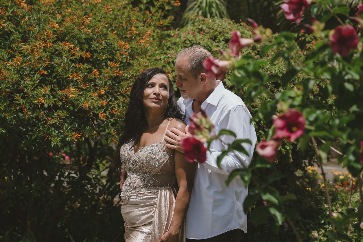 Couple embracing in a lush garden with red flowers, woman in gold dress, man in white shirt.