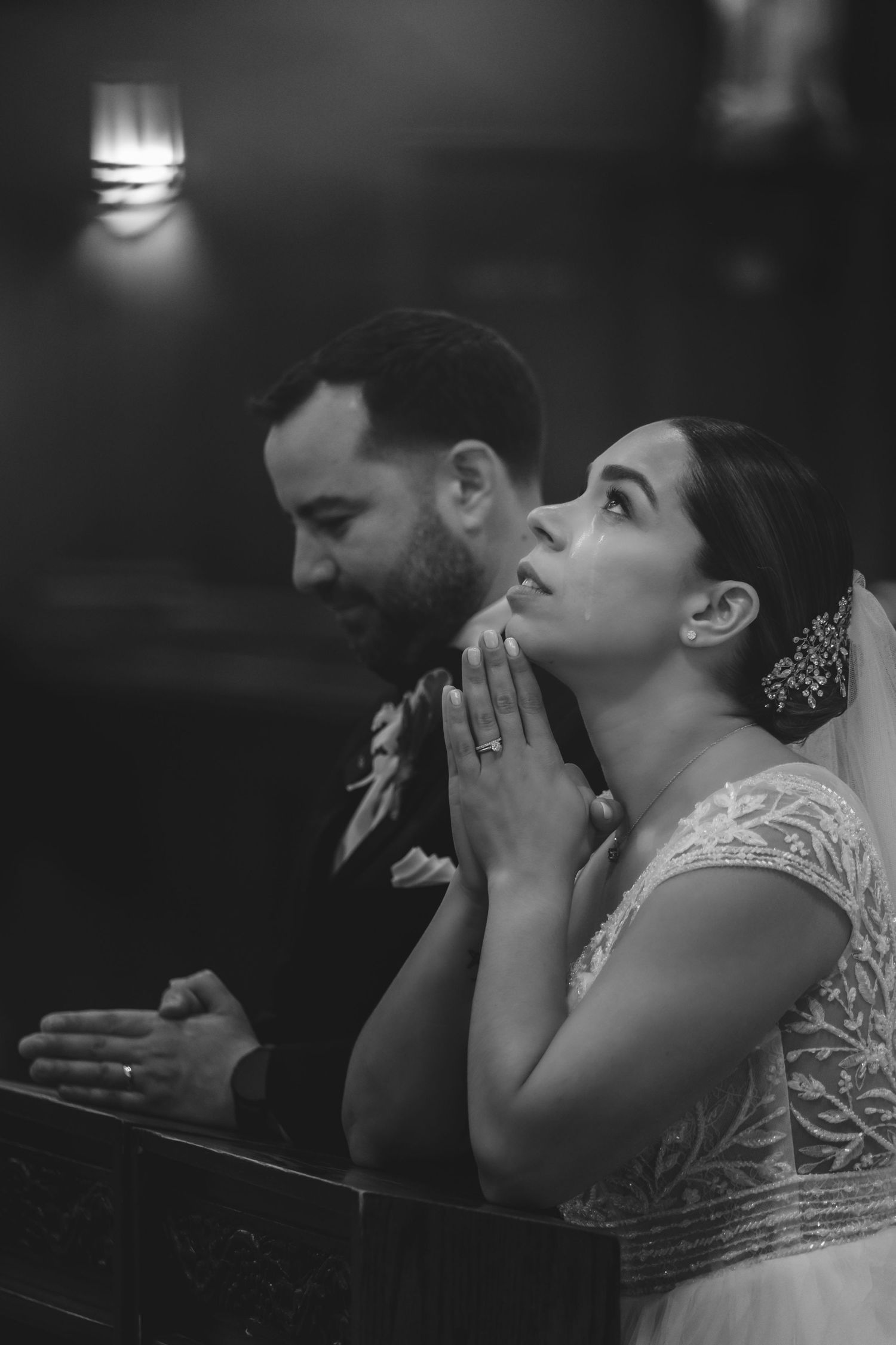 Black and white photo of a bride praying with hands clasped beside her groom during a church wedding ceremony.
