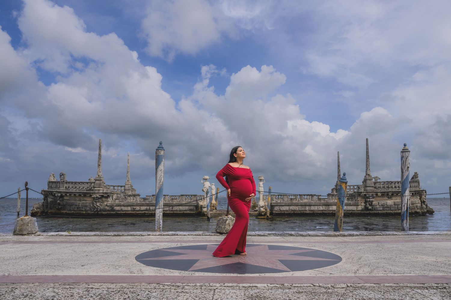 Pregnant woman in red gown poses at waterfront monument under dramatic cloudy sky.