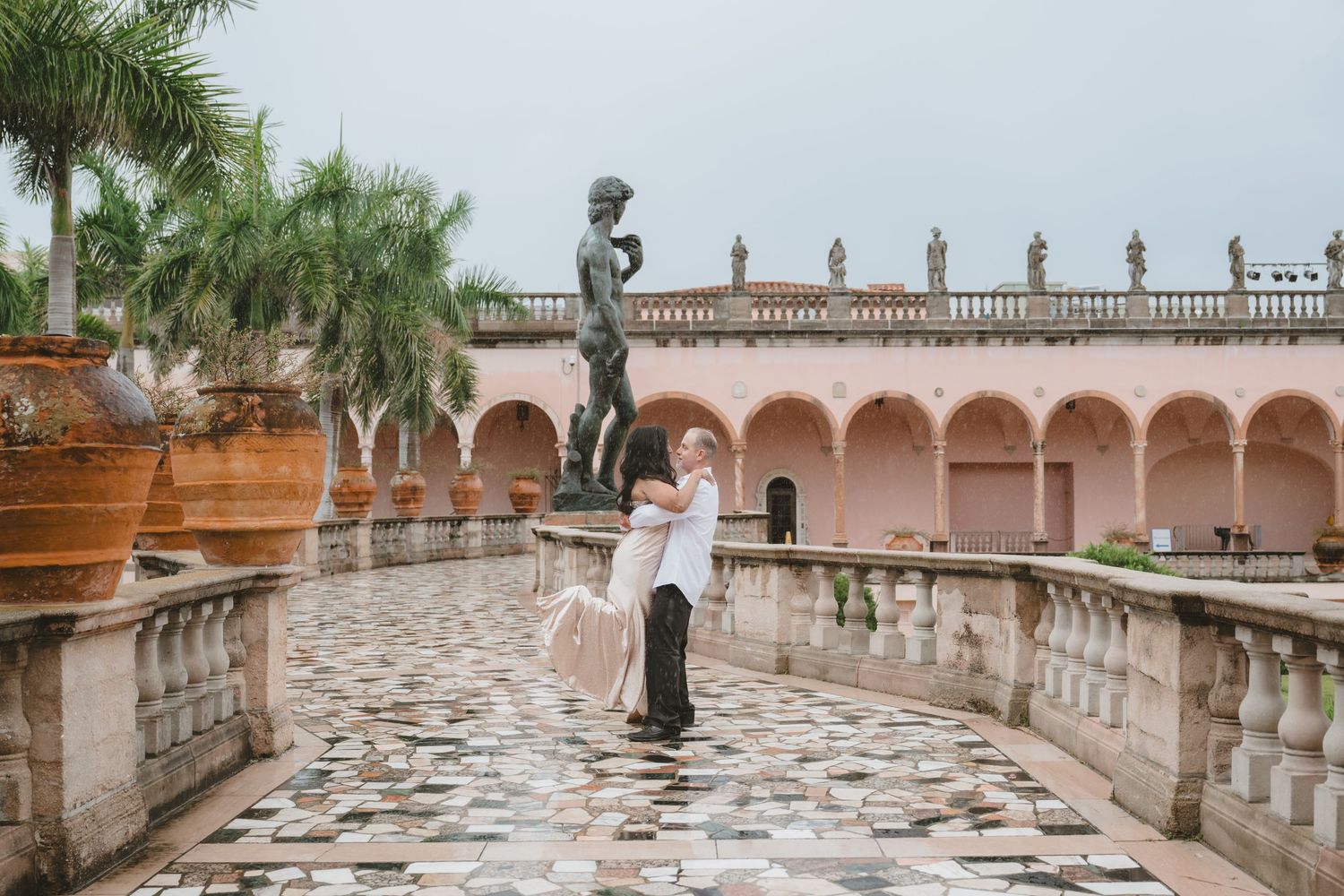 Couple embracing at the Ringling Museum courtyard in Sarasota, FL, with a bronze statue and pink colonnade.