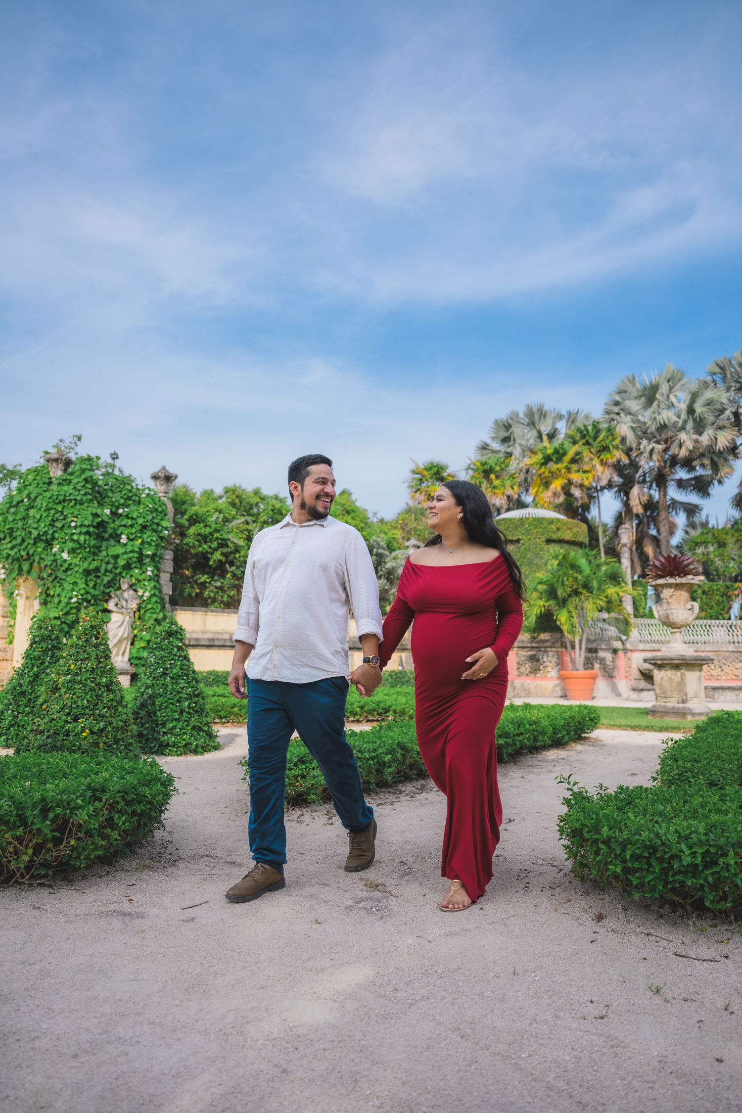 Pregnant couple holding hands and walking in a lush garden, woman in red maternity gown, man in white shirt.