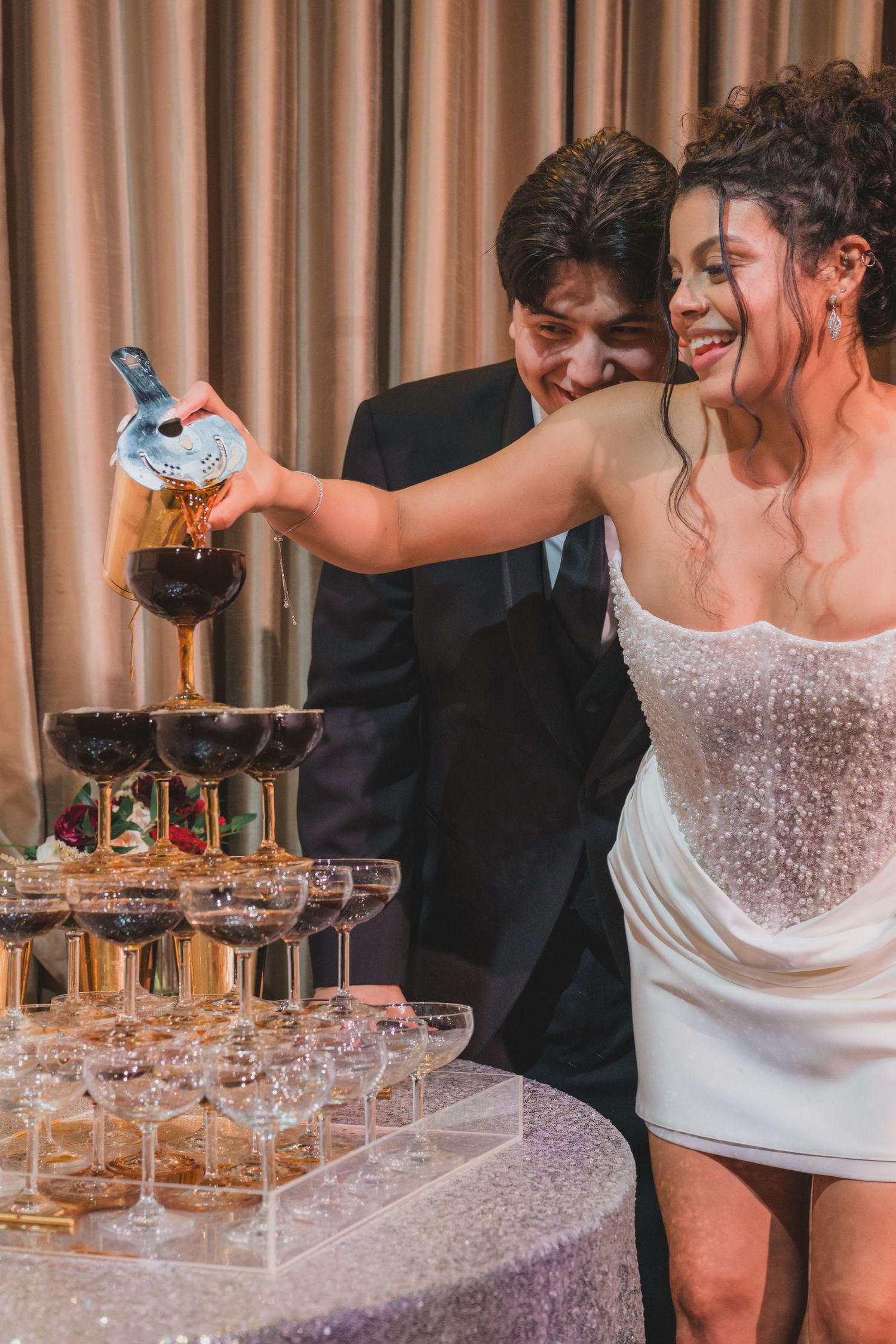Bride and groom pouring champagne tower at wedding reception, smiling couple in white dress and black suit.