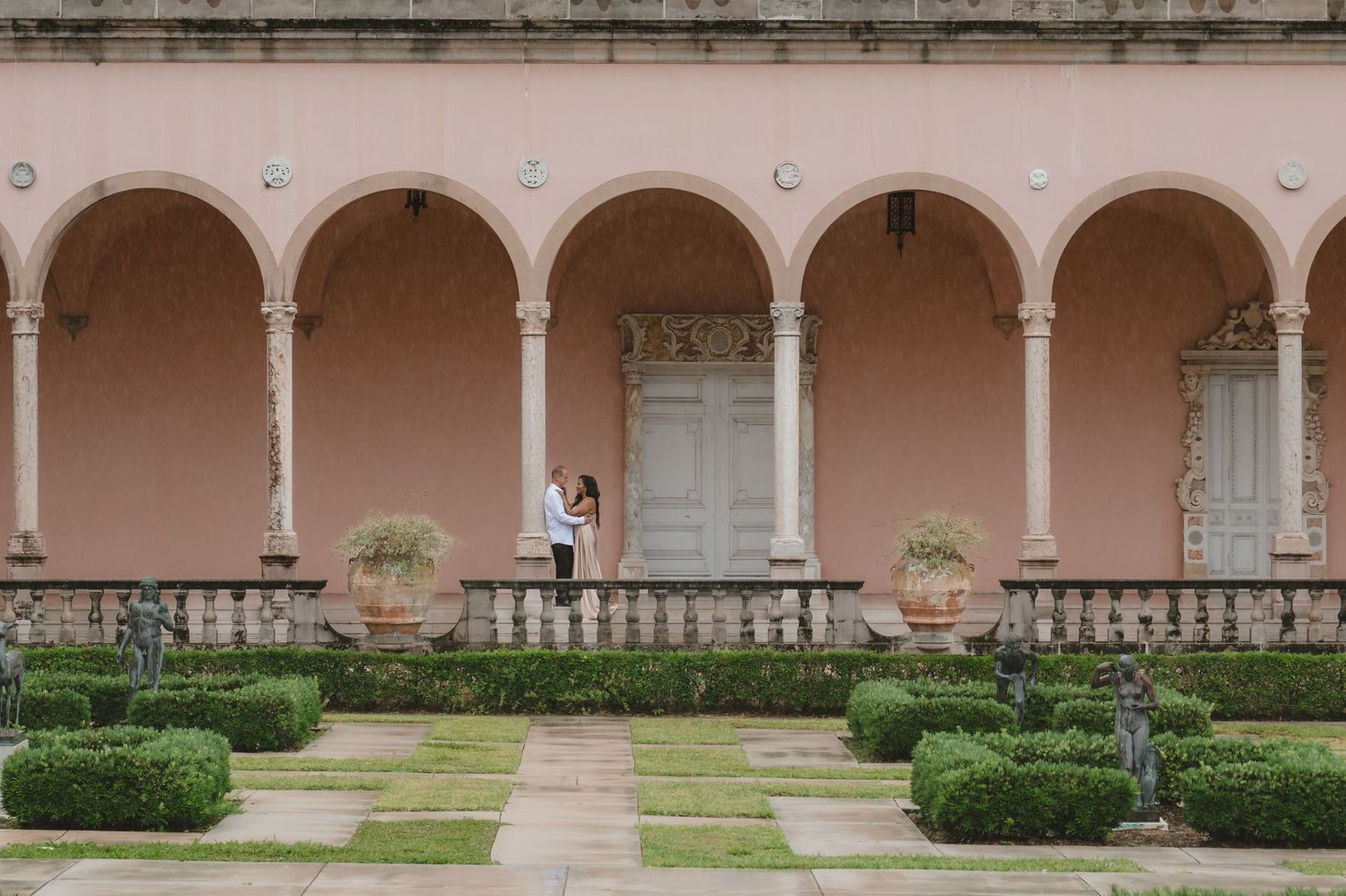 Couple embracing under arched colonnade of elegant pink Italian villa with manicured hedges and garden statues.