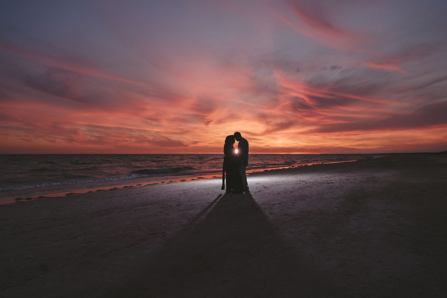 Romantic silhouette of couple kissing on beach during vibrant pink and purple sunset by the ocean.