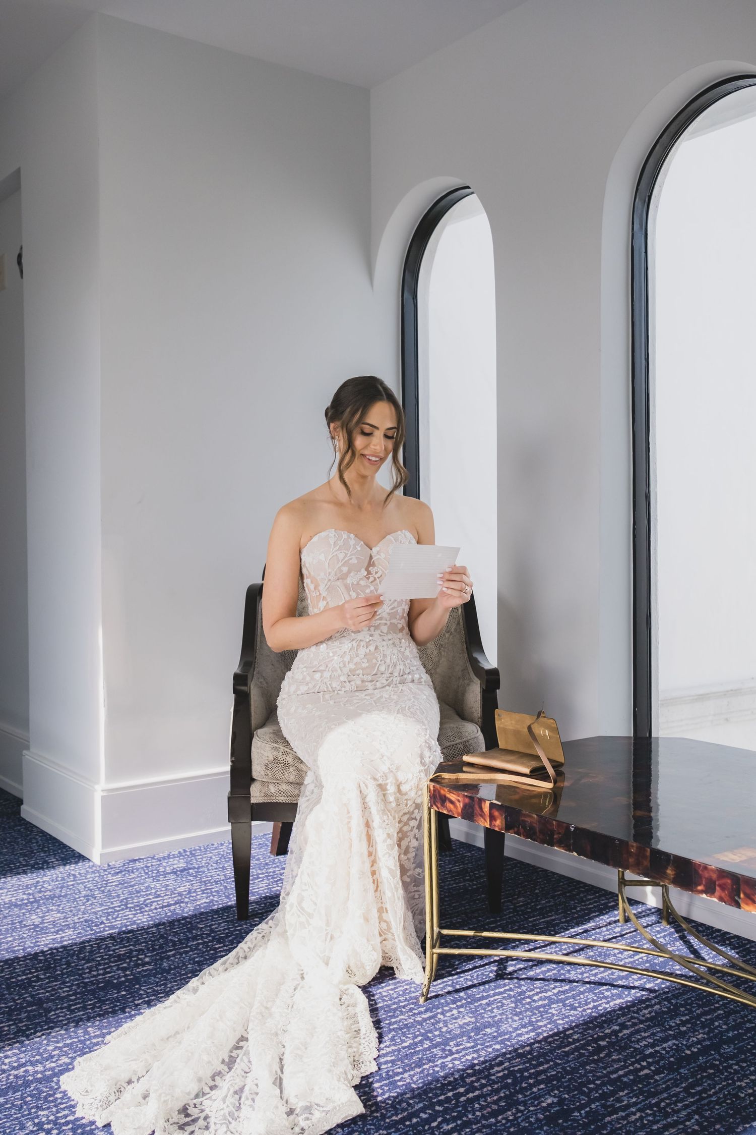 Brunette bride in lace mermaid gown sitting in chair reading letter near arched mirror.