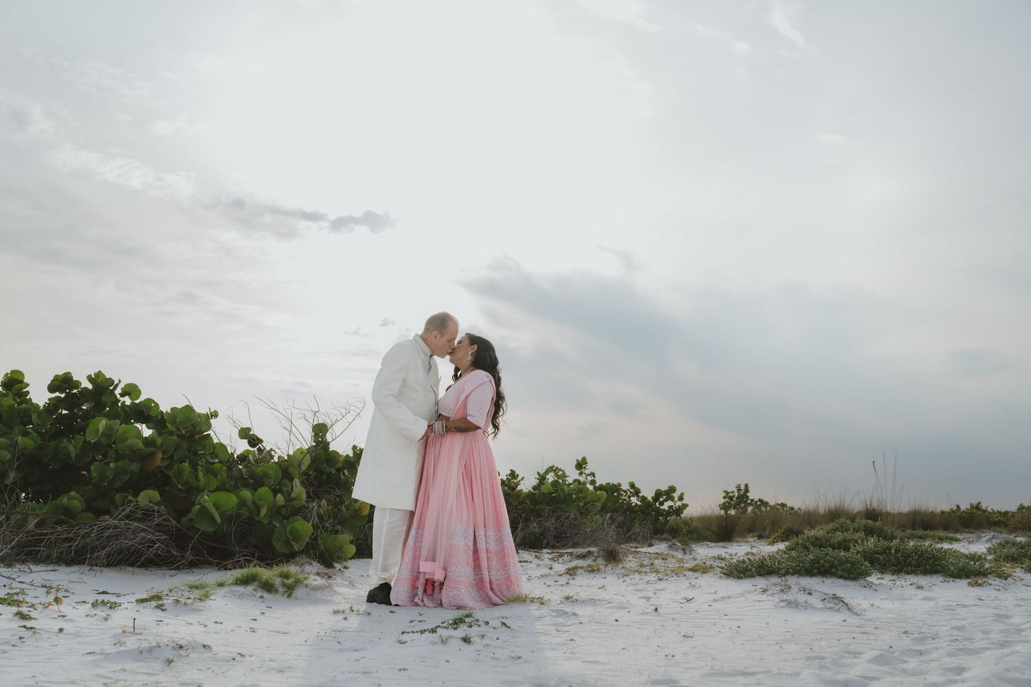 Couple sharing a romantic kiss on white sand beach at sunset, man in white suit and woman in pink gown.