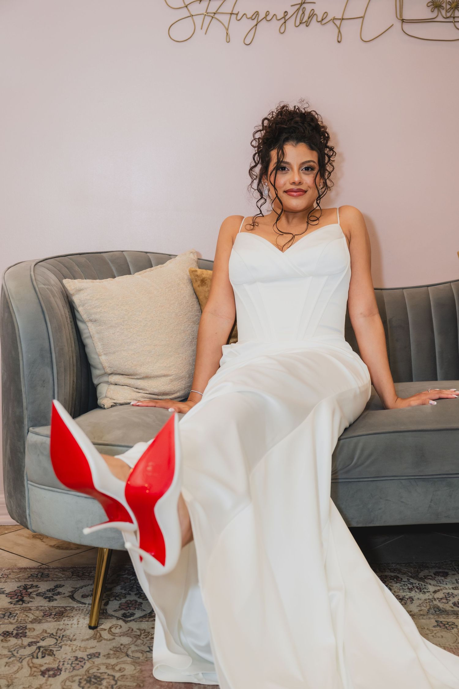 Bride in white gown seated on gray sofa showing red Louboutin heels with curly updo hairstyle.