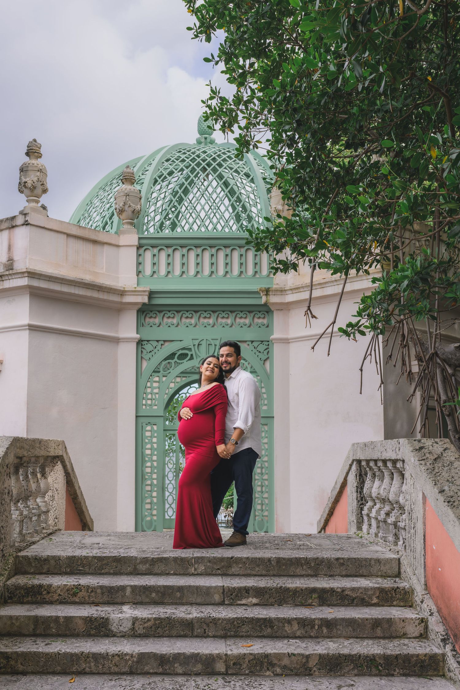 Couple embracing on steps before green latticed dome pavilion, woman in red maternity gown.