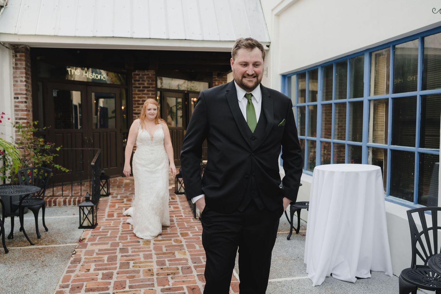Groom in black suit with green tie walks ahead of bride on brick pathway before ceremony.