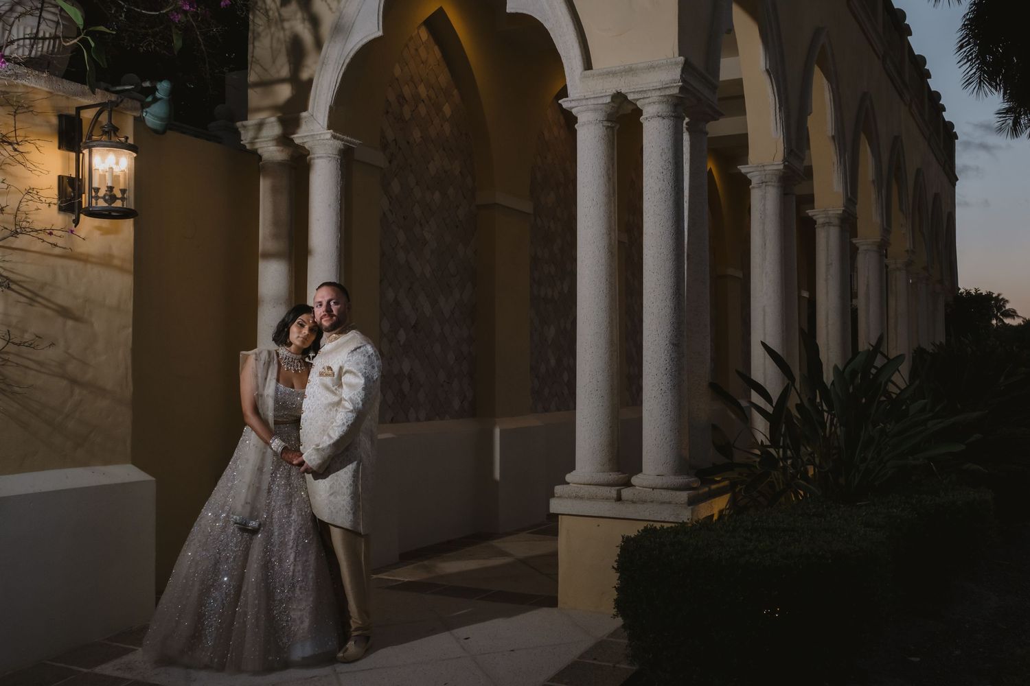 Indian bride and groom in traditional attire sharing intimate moment under arched colonnade at dusk.