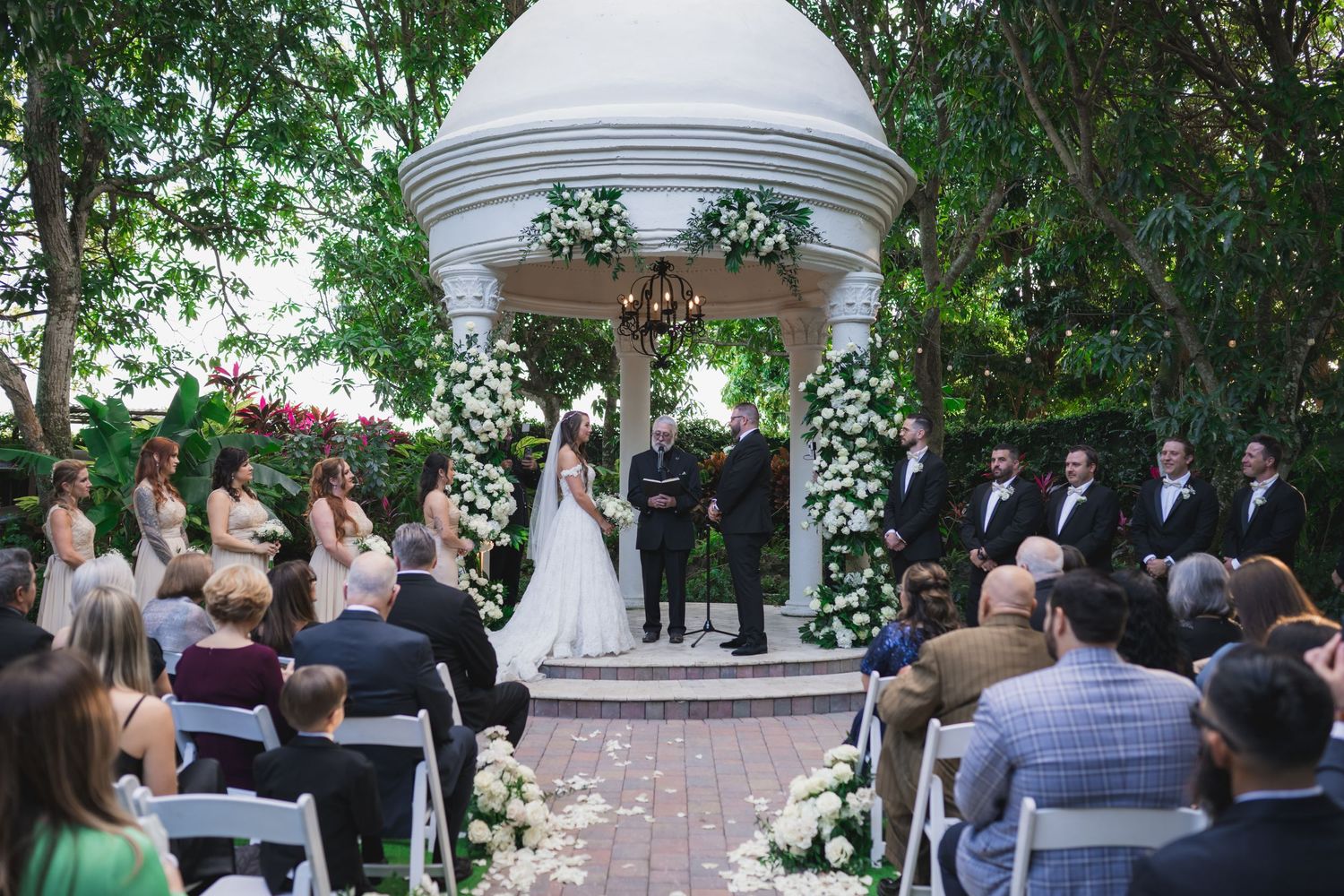 Elegant outdoor wedding ceremony under a white gazebo adorned with white flowers and lush greenery.