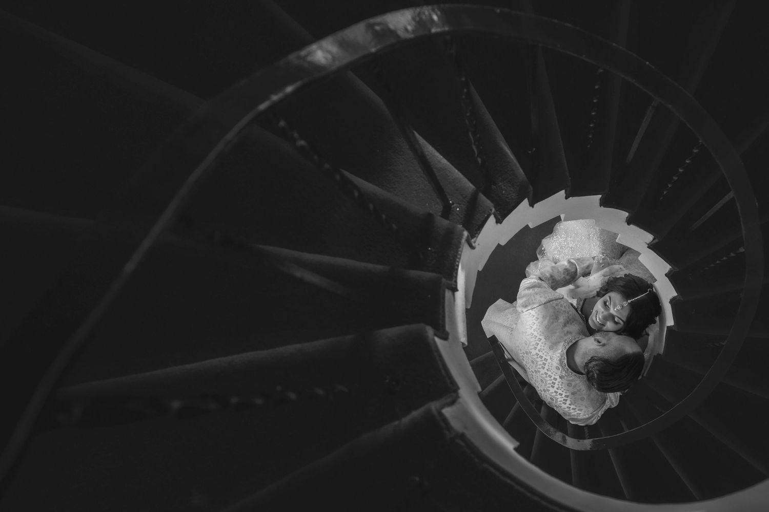 Black and white aerial view of a couple embracing at the bottom of an elegant spiral staircase.