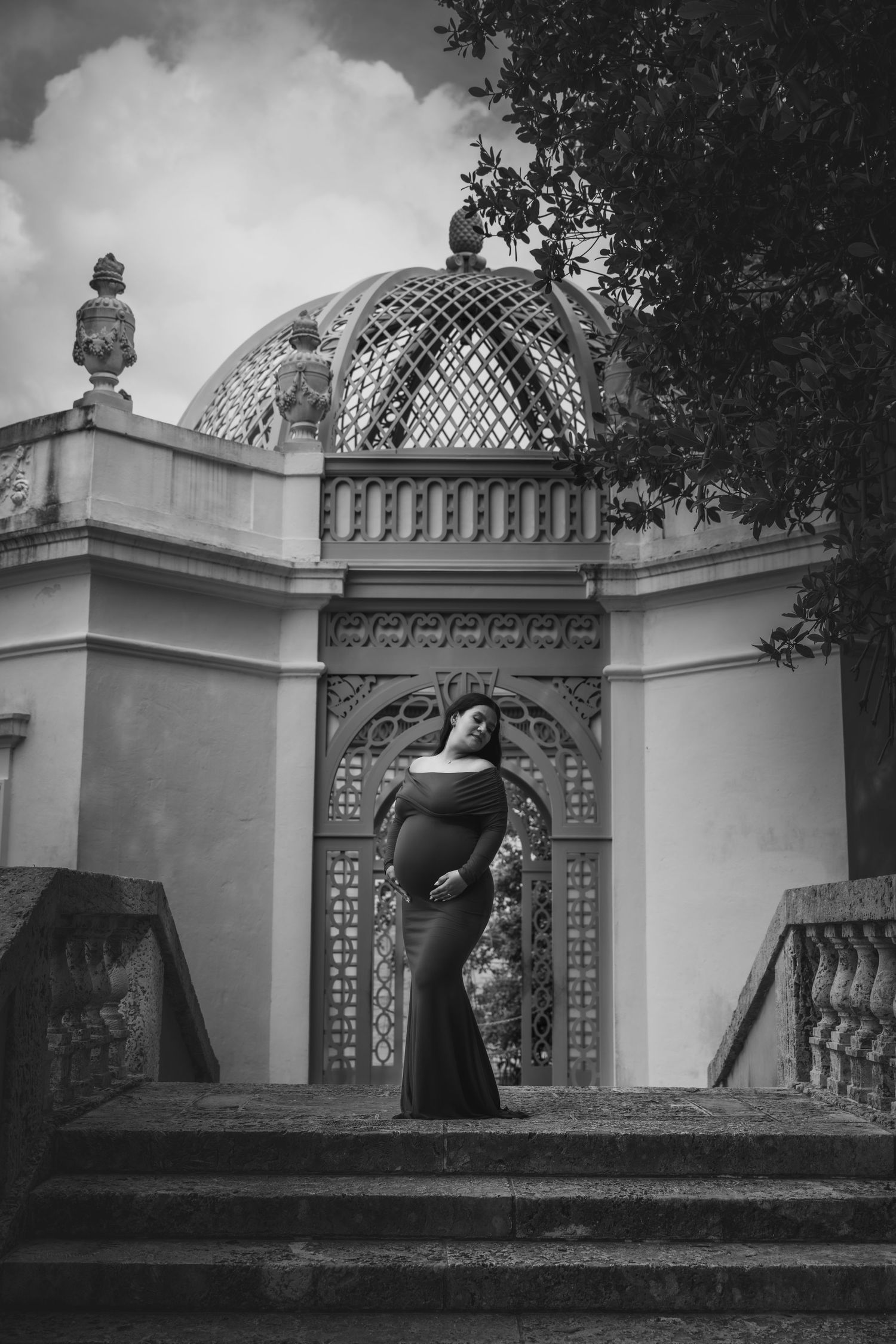 Pregnant woman in black gown poses on stone steps before ornate domed pavilion in black and white photo.