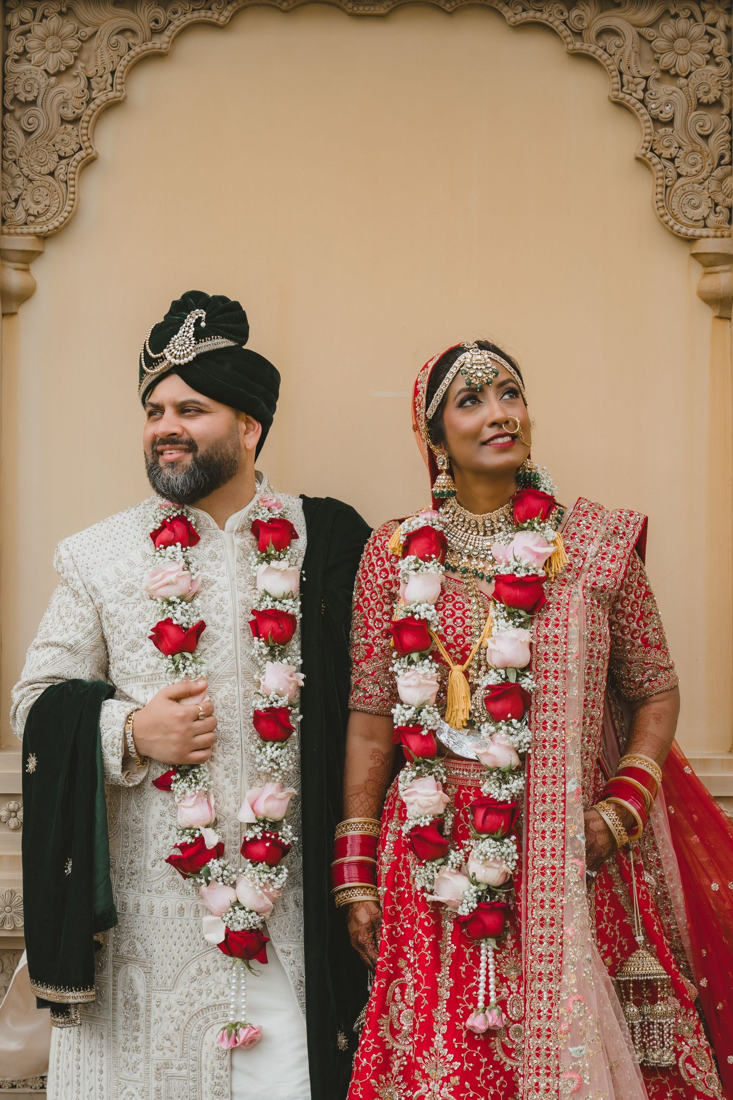 Indian bride in red lehenga and groom in white sherwani wearing floral garlands at ceremony.