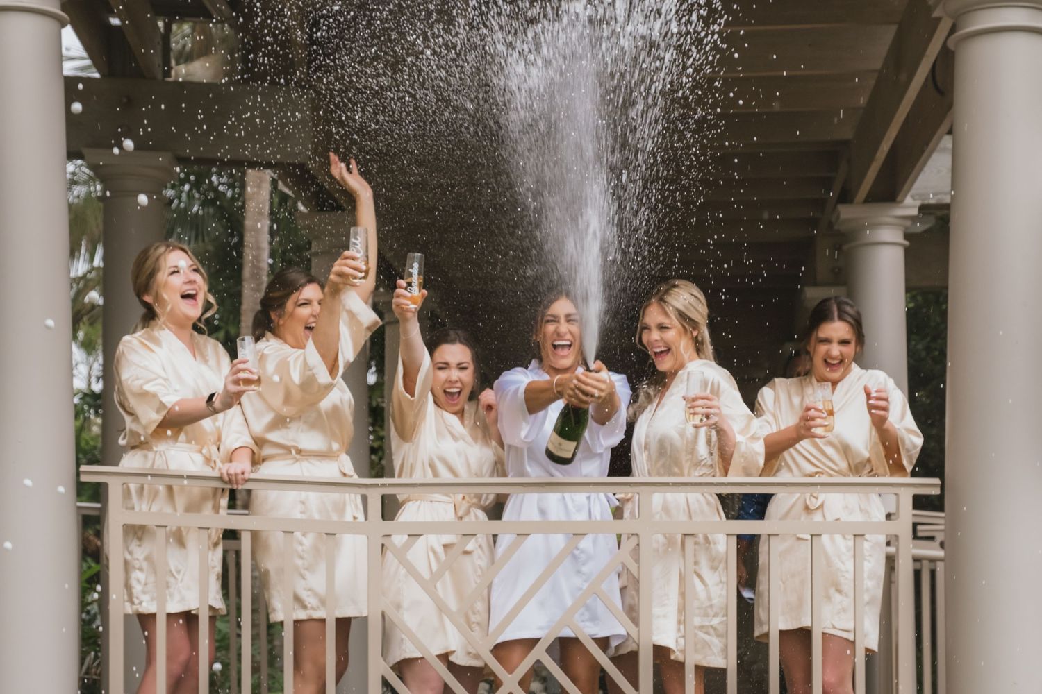 Bridesmaids in champagne robes celebrate with a champagne spray on a balcony at a wedding.