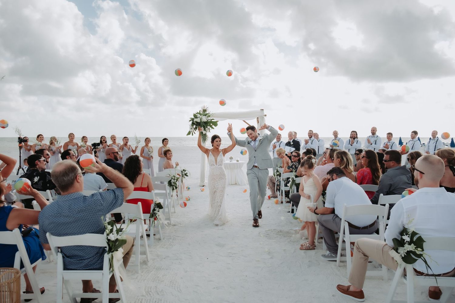 Bride and groom celebrate at beach ceremony aisle as balloons float into cloudy sky.