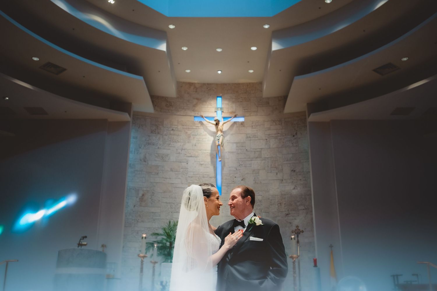 Bride and groom share a tender kiss beneath a crucifix in a modern Catholic church.