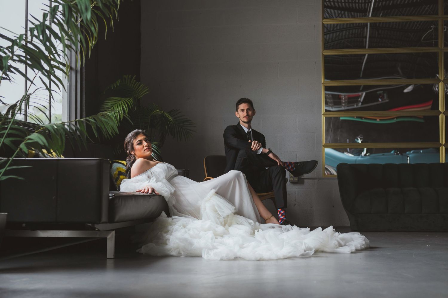 Bride in white gown reclines on sofa while groom in black suit sits behind her in modern interior.