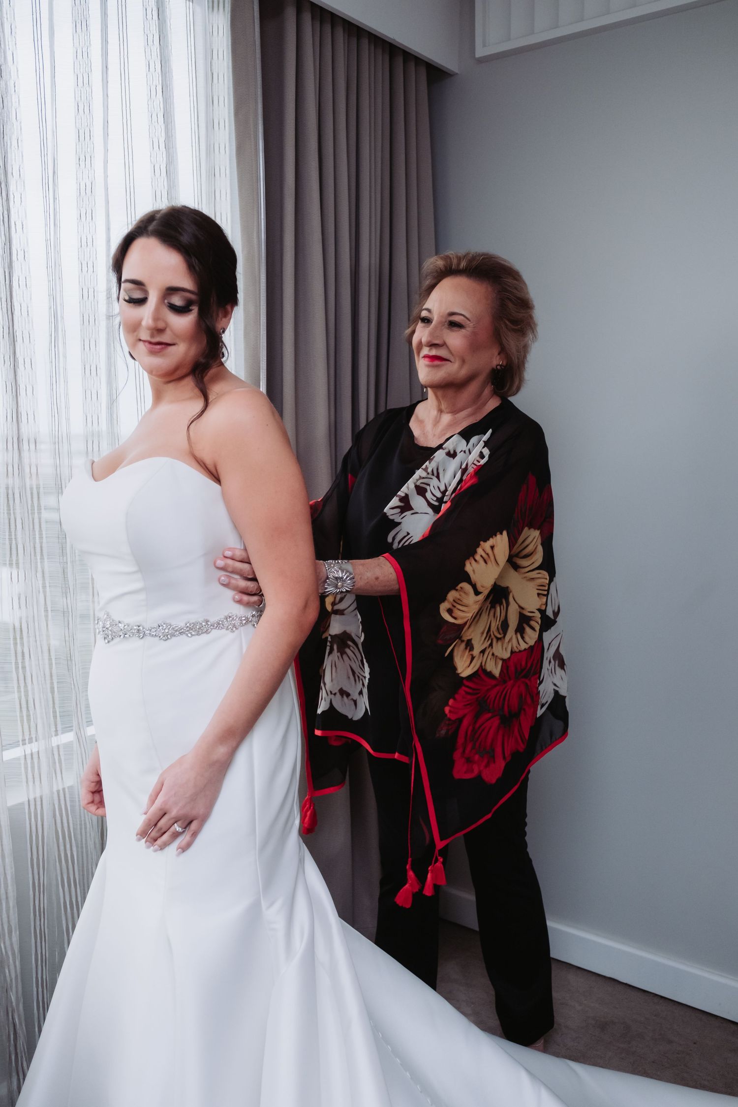 Older woman helping bride zip up white strapless wedding gown near window with gray curtains.