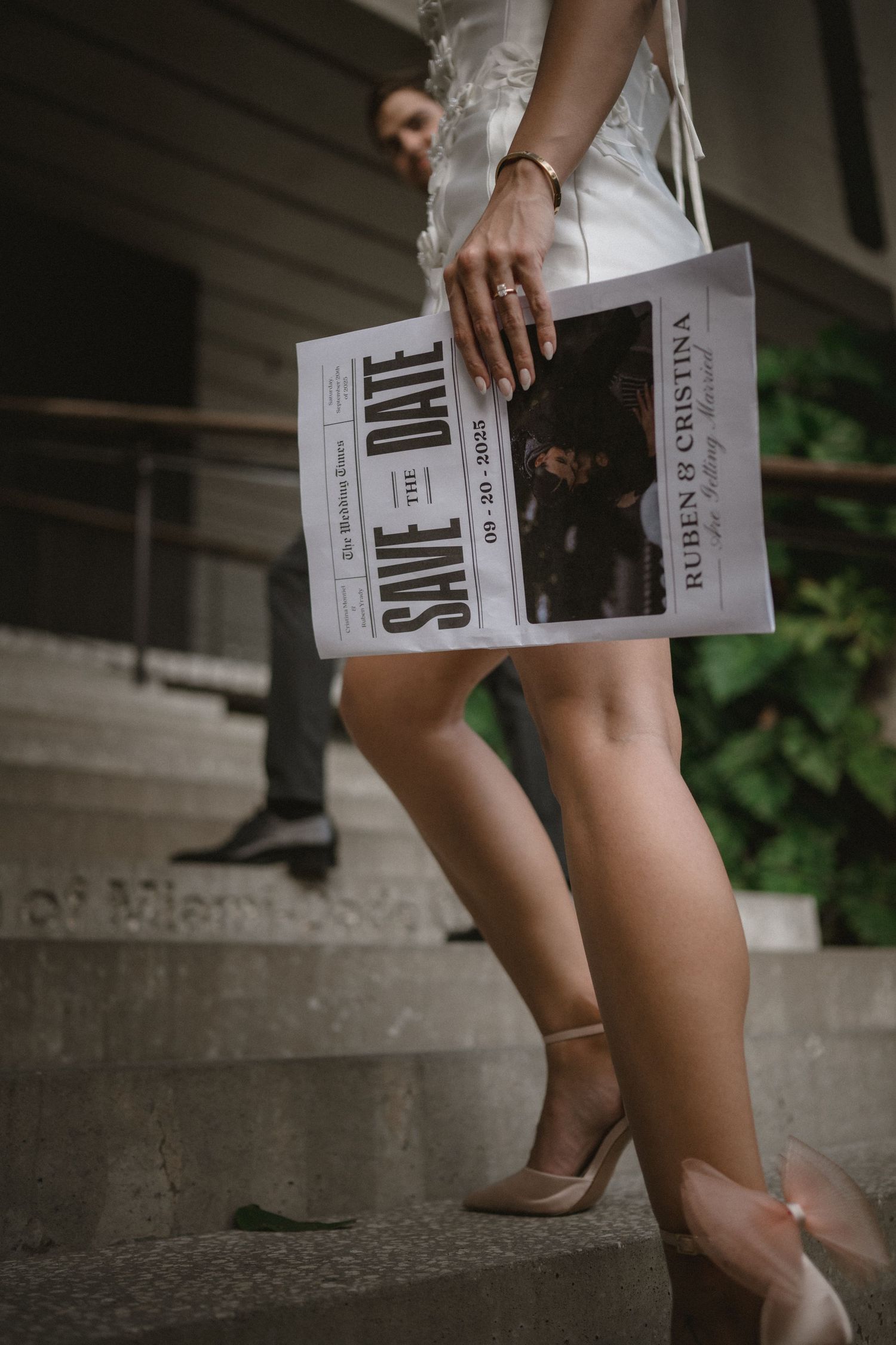 Bride holding a Save the Date newspaper announcement for Ruben and Cristina while descending steps.