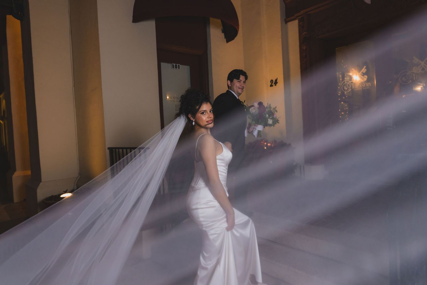 Bride in white gown with flowing veil stands beside groom in elegant venue with dramatic light rays.