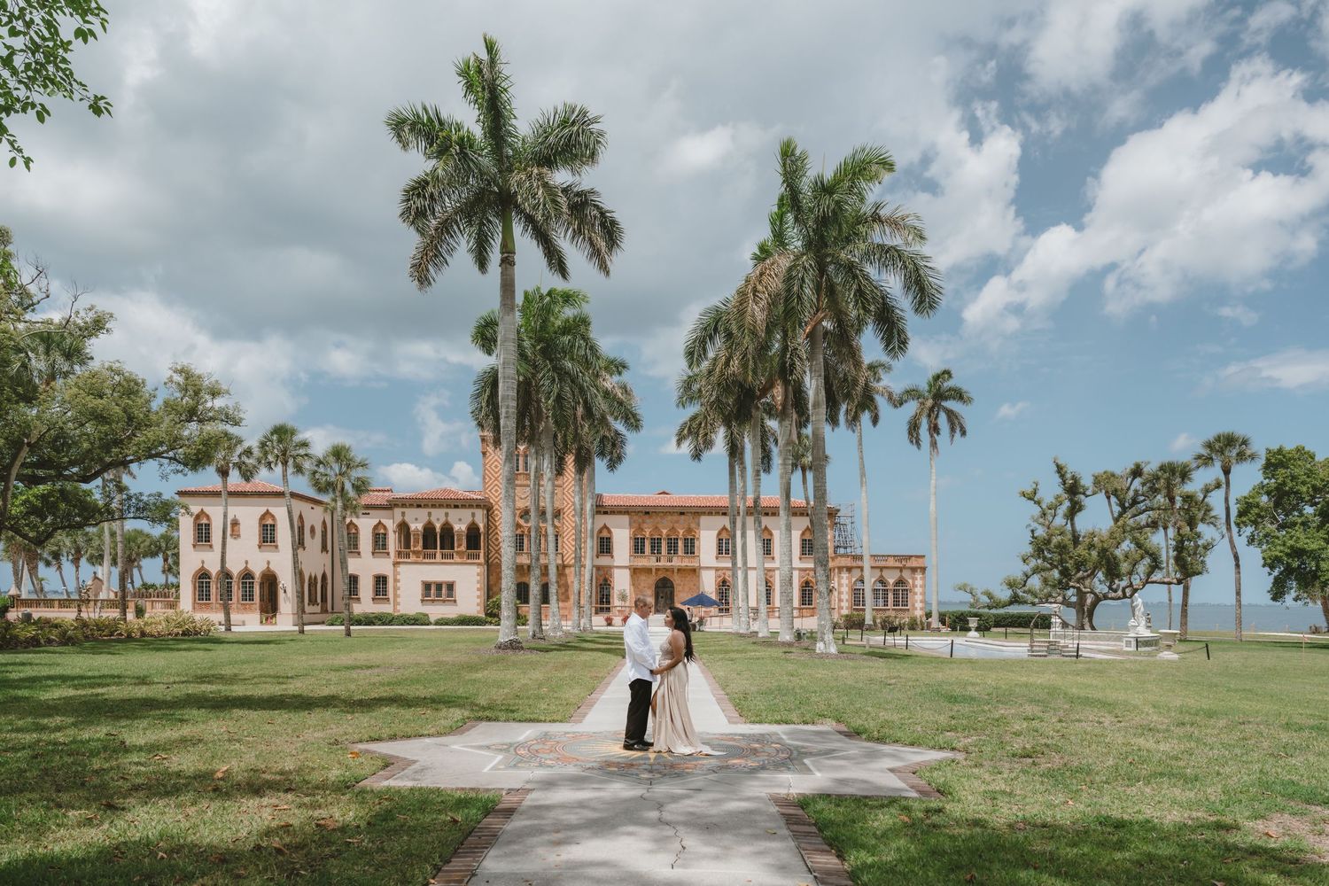 Couple embracing on a pathway in front of a grand historic estate surrounded by tall palm trees in Florida.