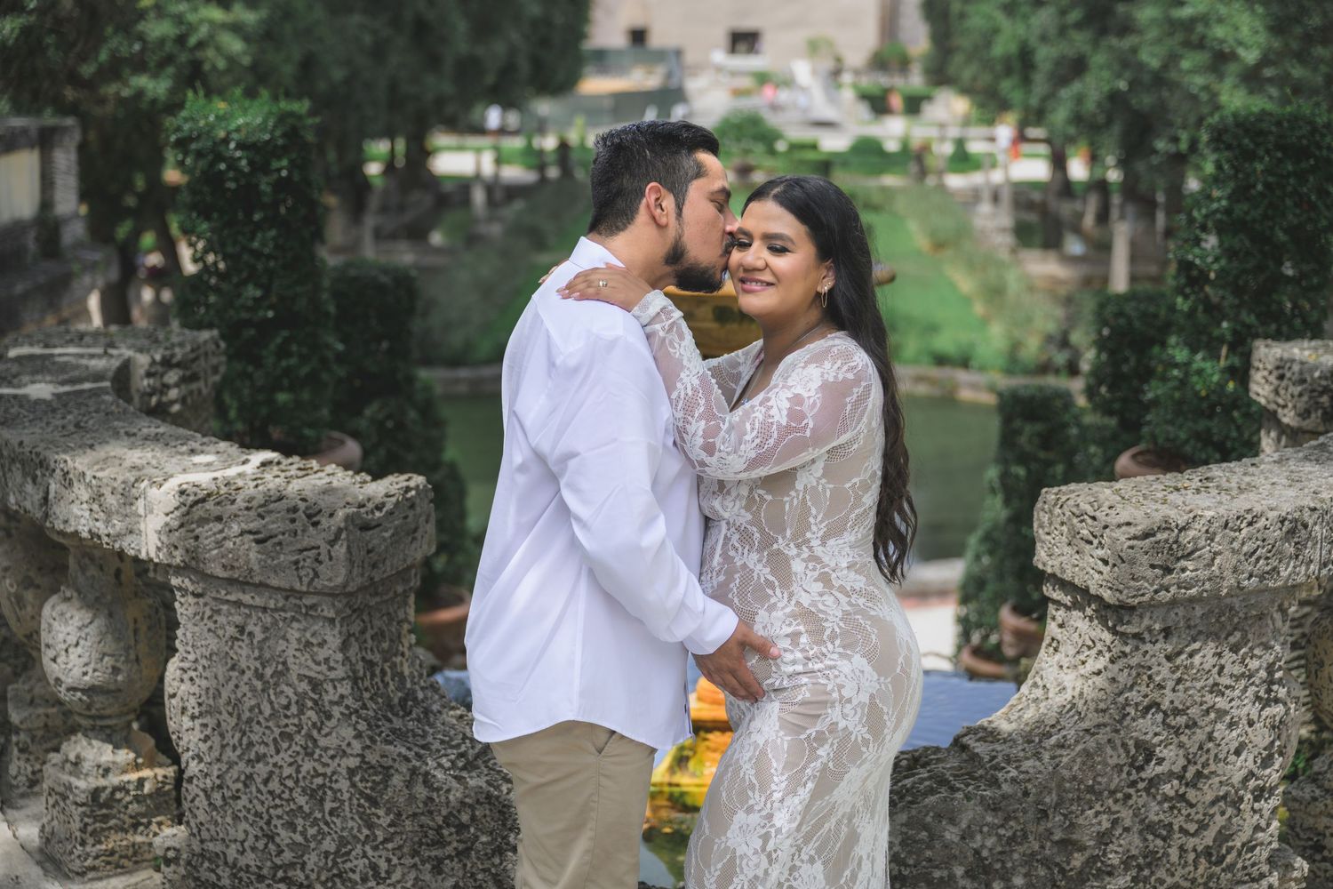 Couple sharing a romantic kiss on a stone balcony surrounded by lush greenery and a serene pond.