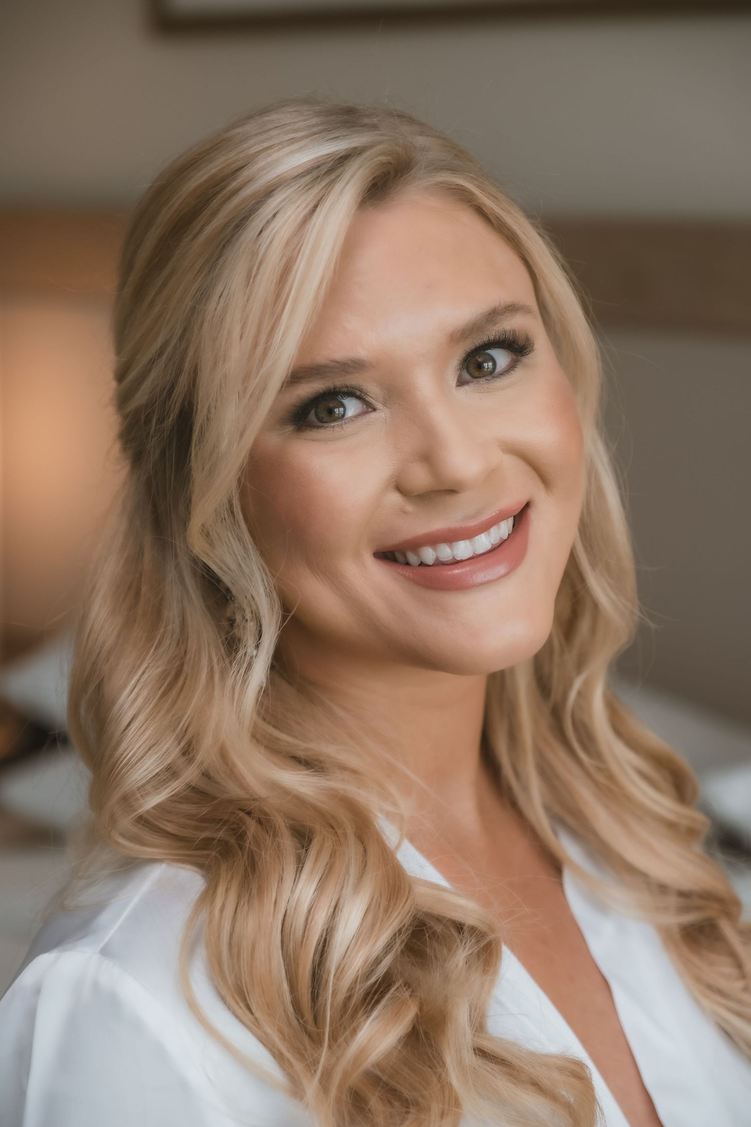 Smiling blonde bride with soft waves and elegant makeup wearing white robe before wedding.