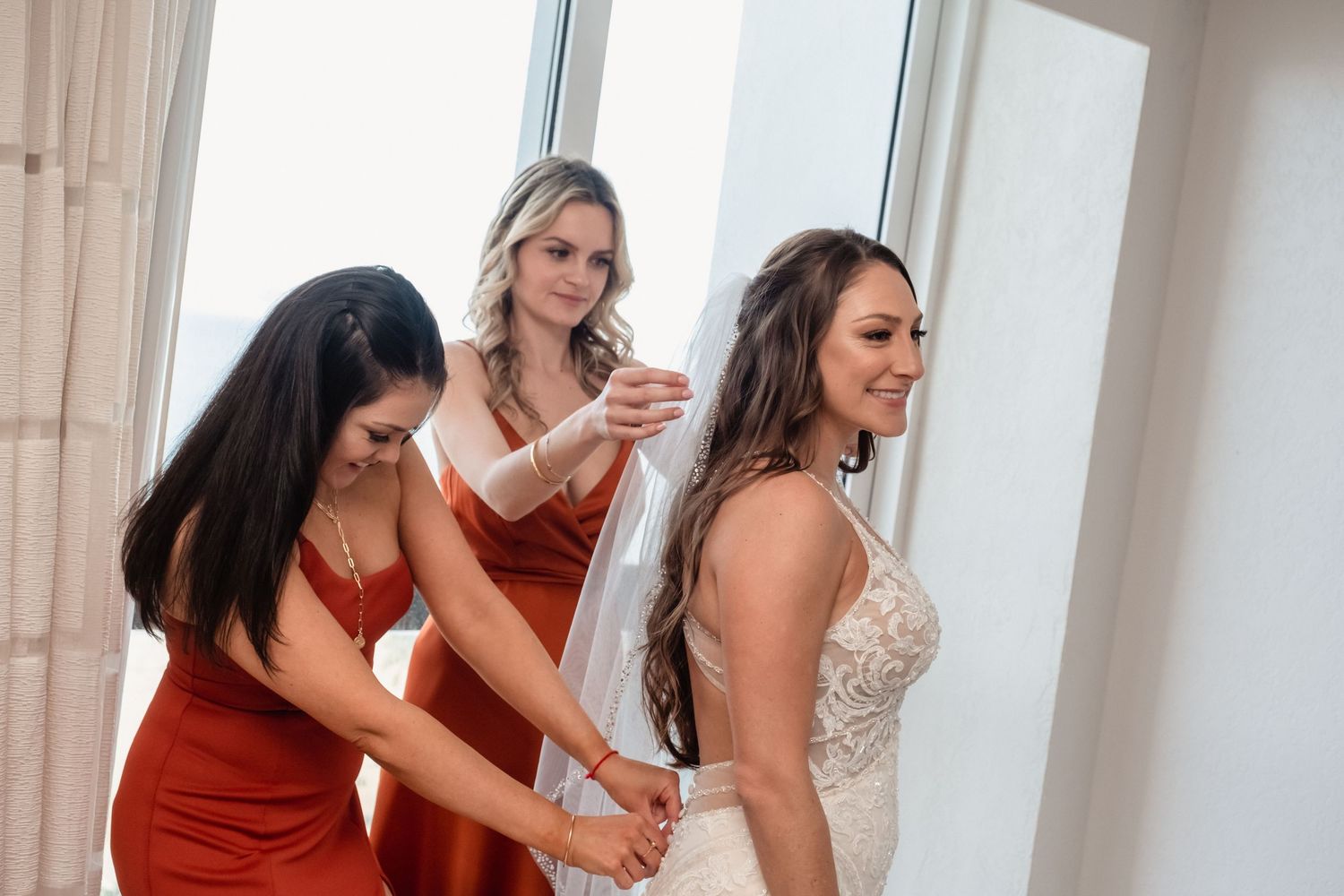 Two bridesmaids in rust satin dresses helping bride with wedding gown near bright window.