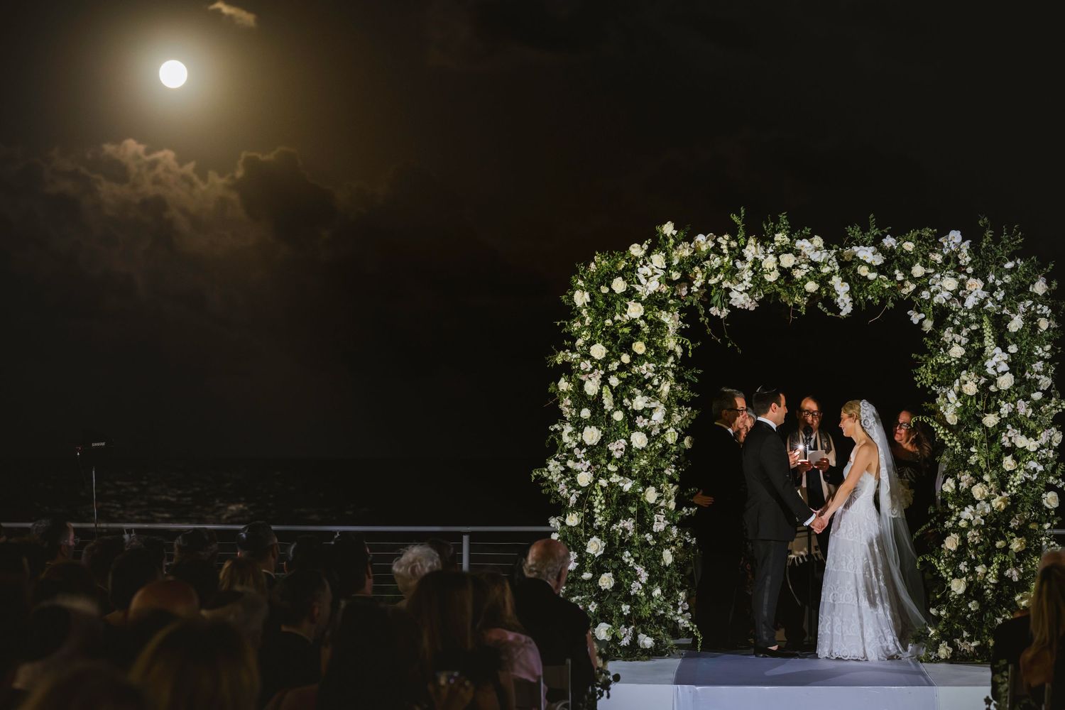 Bride and groom exchange vows under a stunning floral arch beneath a full moon at night.