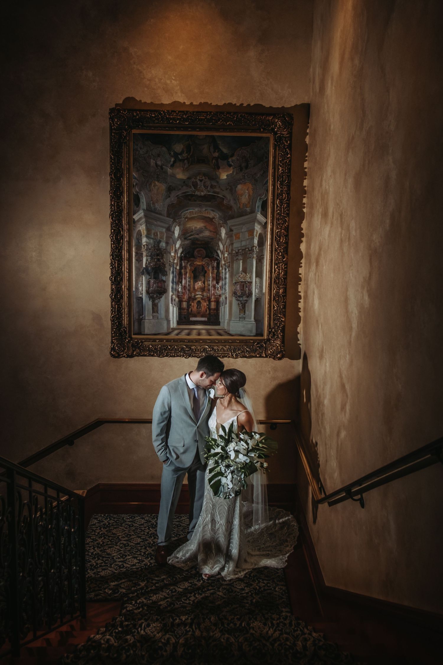 Bride and groom sharing an intimate moment on a staircase beneath a grand ornate painting in a historic venue.