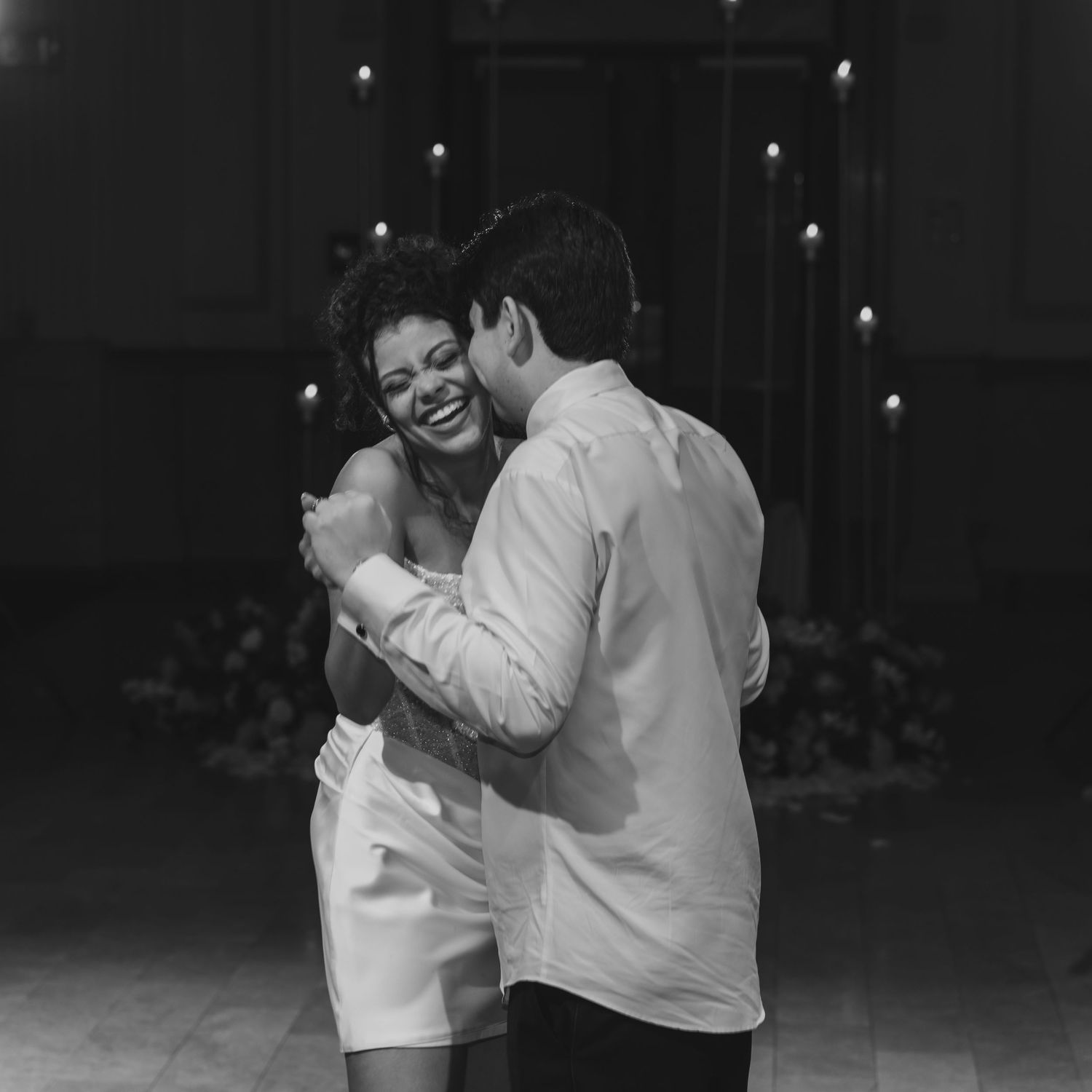 Black and white photo of a couple sharing a joyful dance, the woman laughing in a white dress at an elegant venue.