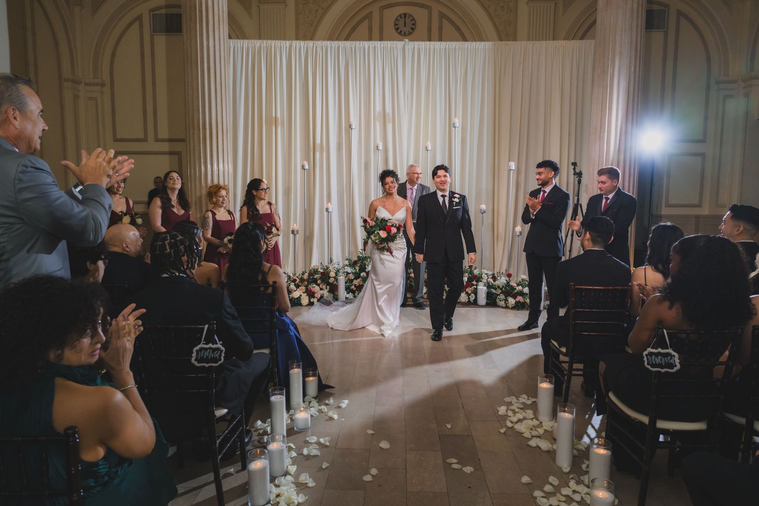 Bride and groom walk down the aisle at indoor wedding ceremony with candles and white floral backdrop.