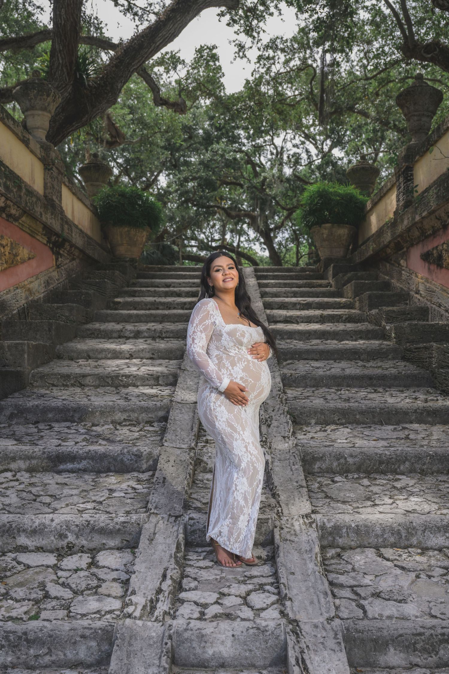Pregnant woman in white lace dress poses on ancient stone staircase surrounded by trees.