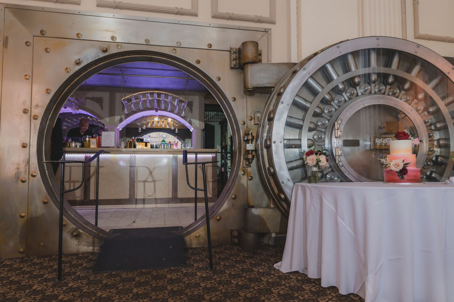 Two large open bank vault doors revealing an elegant bar and event space with purple ambient lighting.