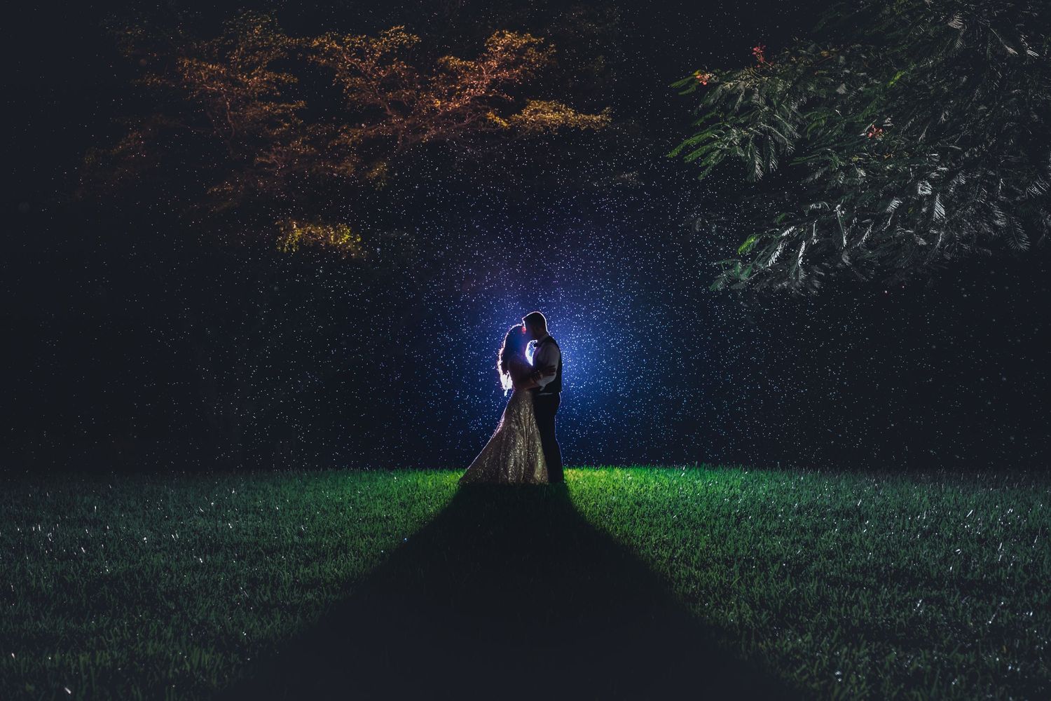 Bride and groom embracing in a glowing light in a dark garden with colorful trees at night.