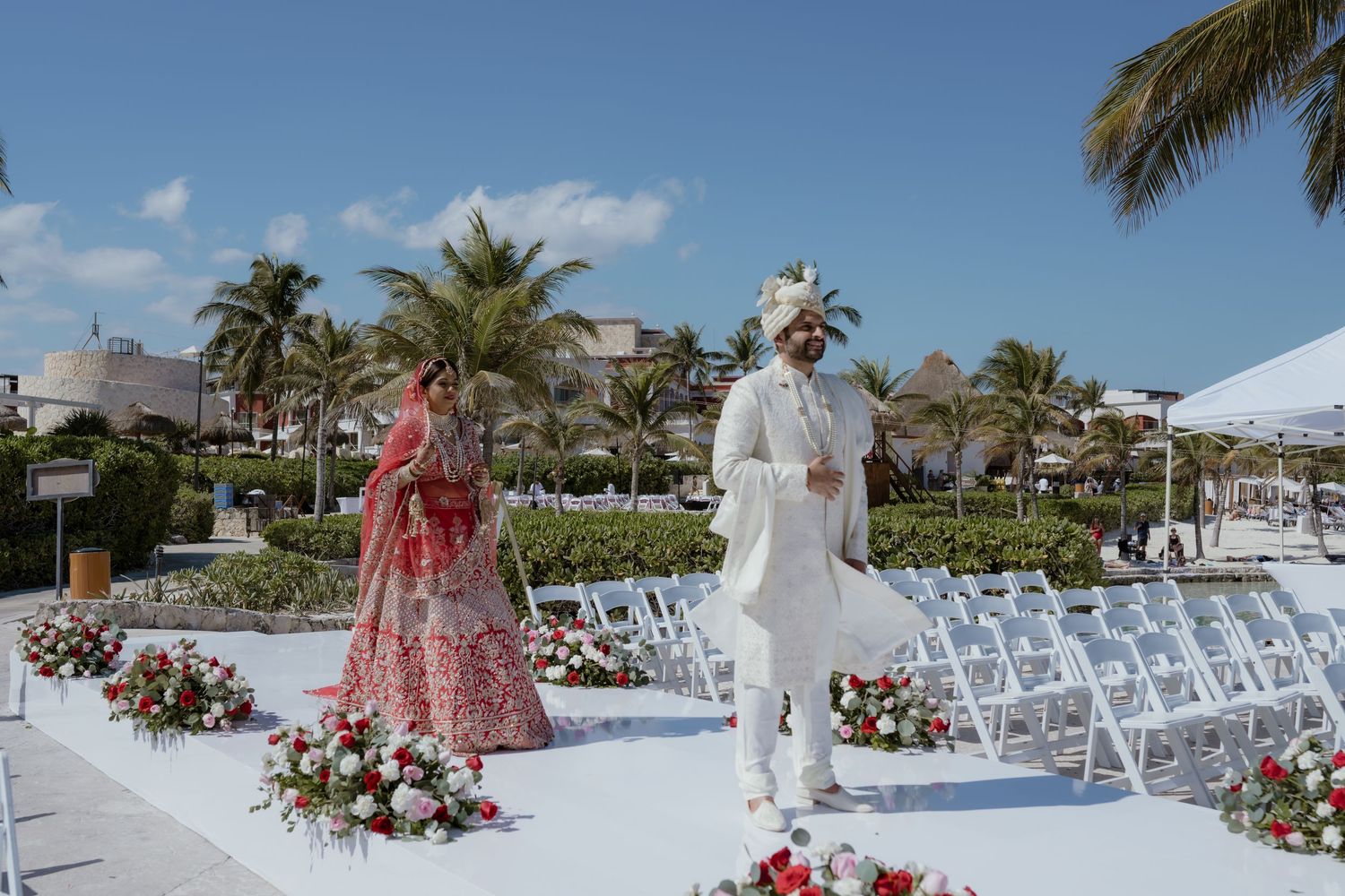 Indian couple in traditional wedding attire walk down outdoor aisle lined with floral arrangements and palm trees.