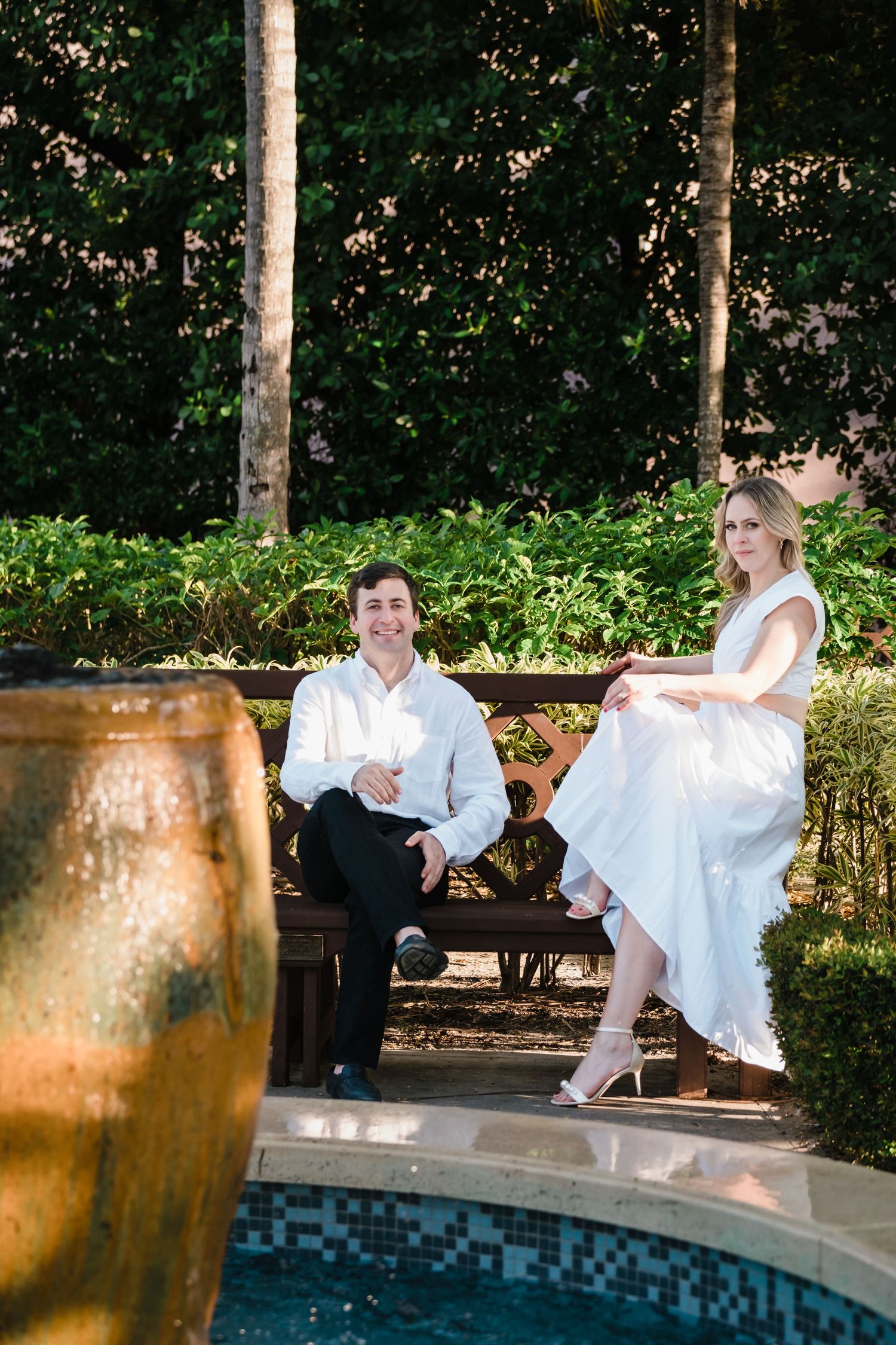 Smiling couple relaxing on garden bench beside decorative fountain in lush park.