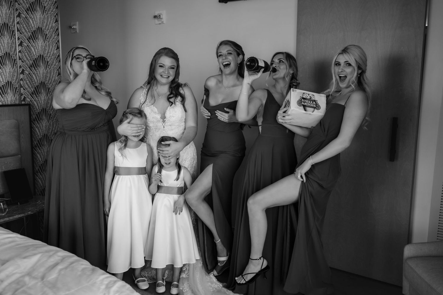 Black and white photo of bridesmaids and flower girls celebrating with drinks in a hotel room.