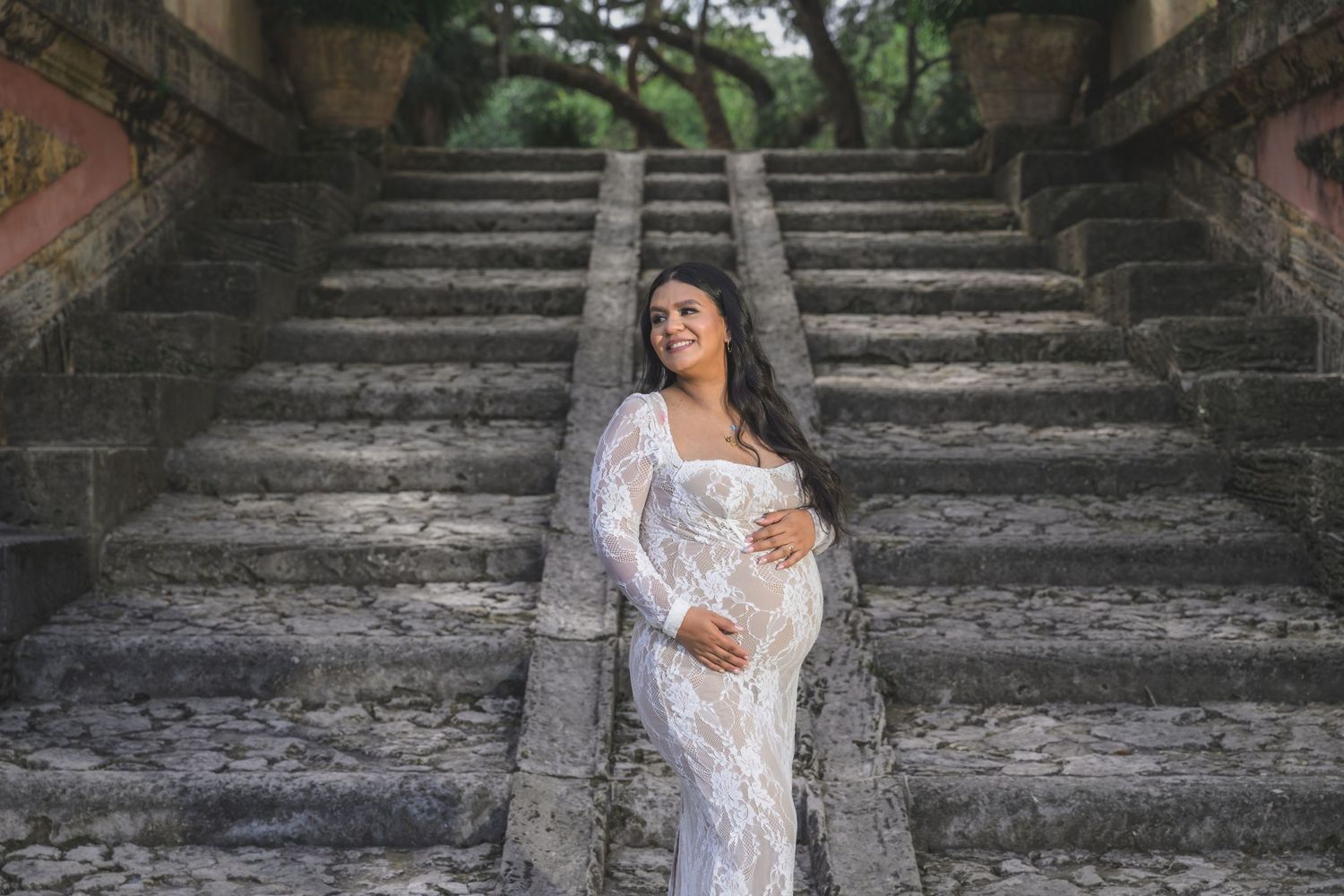 Pregnant woman in white lace gown poses on stone staircase outdoors during maternity photo shoot.