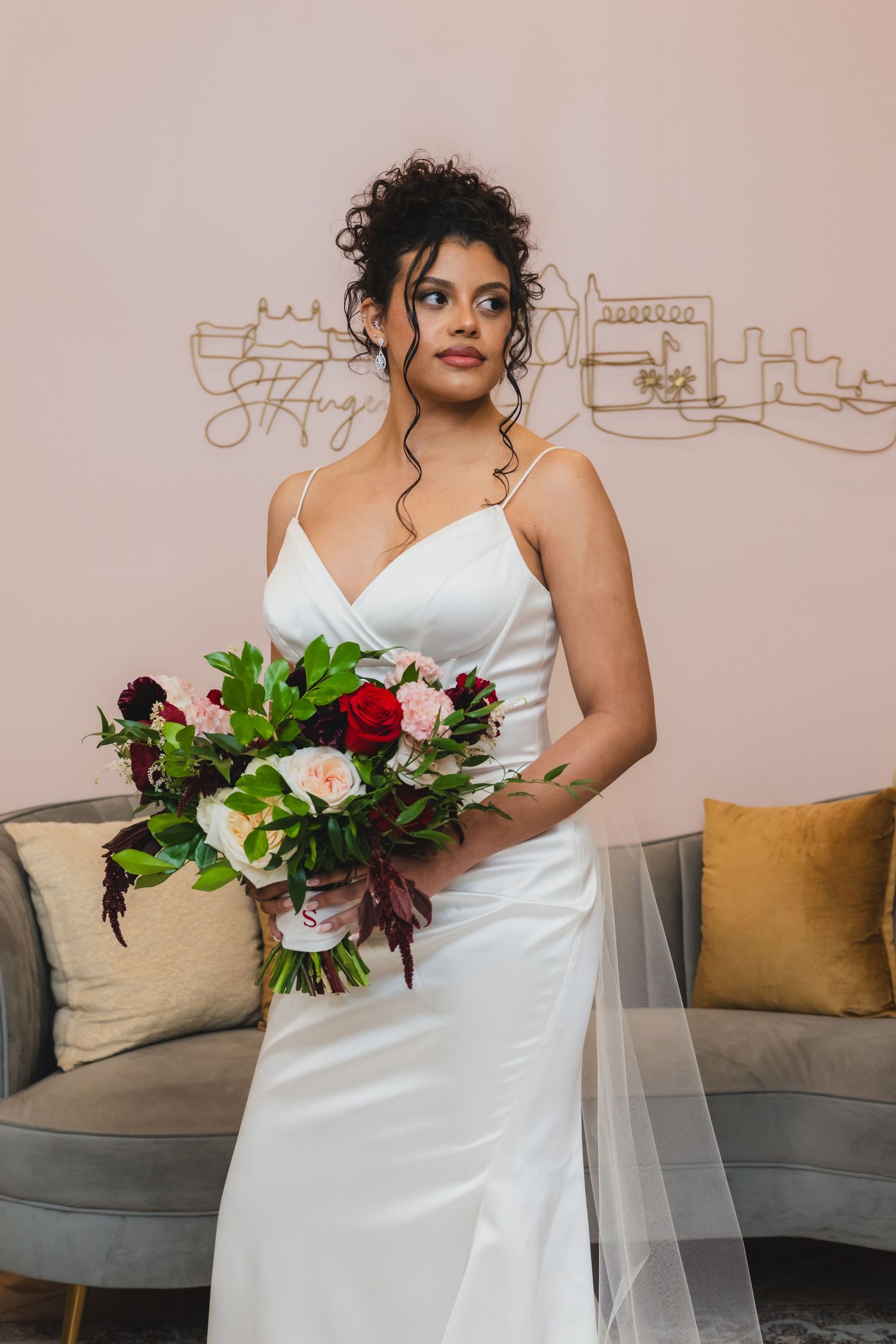 Bride in white satin gown holding red and pink bouquet, standing before a pink wall with St. Augustine skyline art.