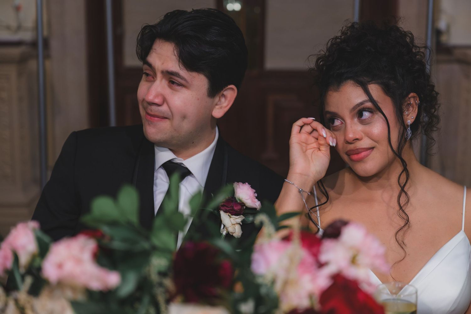Emotional bride and groom crying tears of joy during wedding reception speeches, surrounded by pink and red floral arrangements.