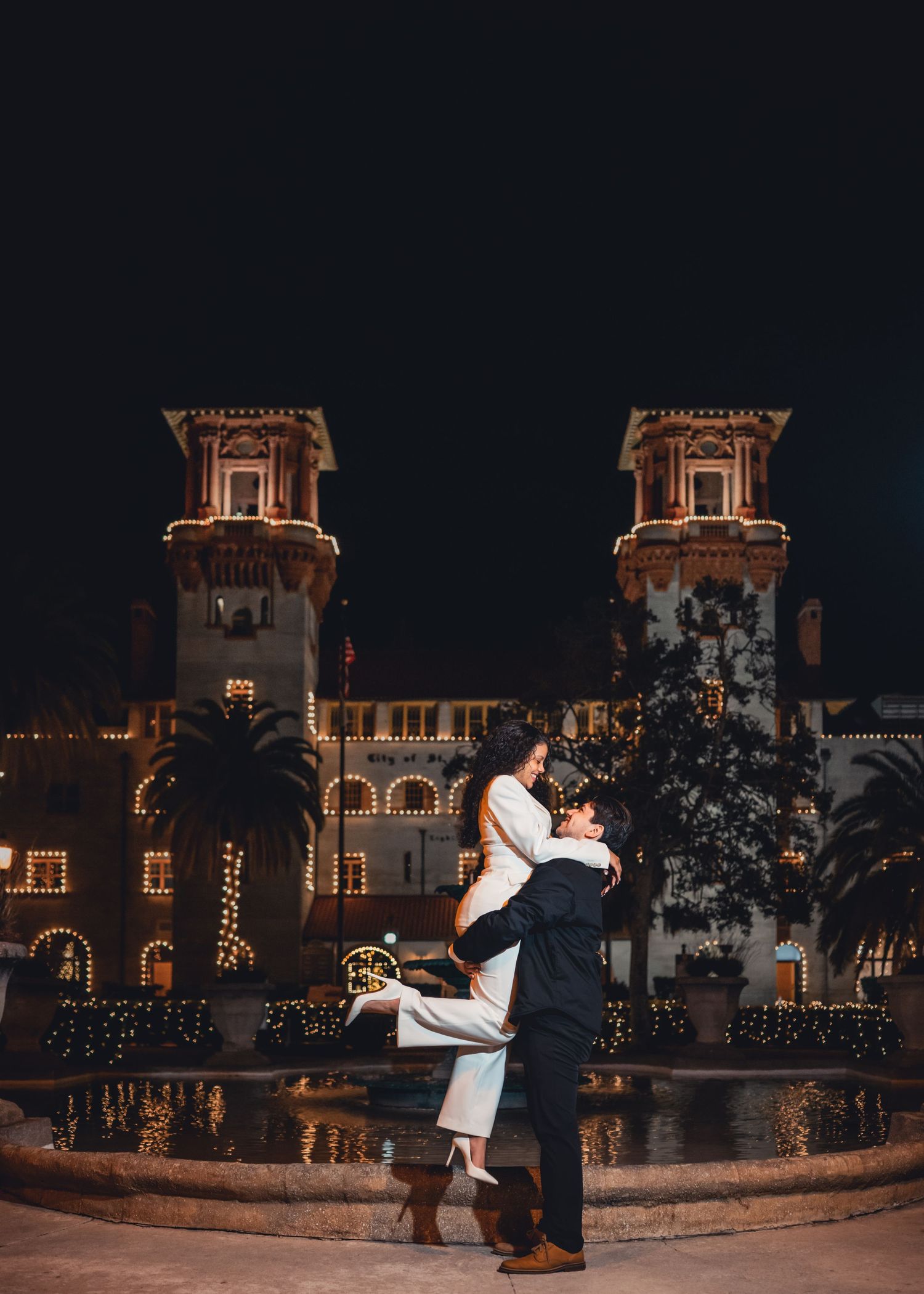 Couple embracing by a fountain at night in front of a beautifully lit historic building.