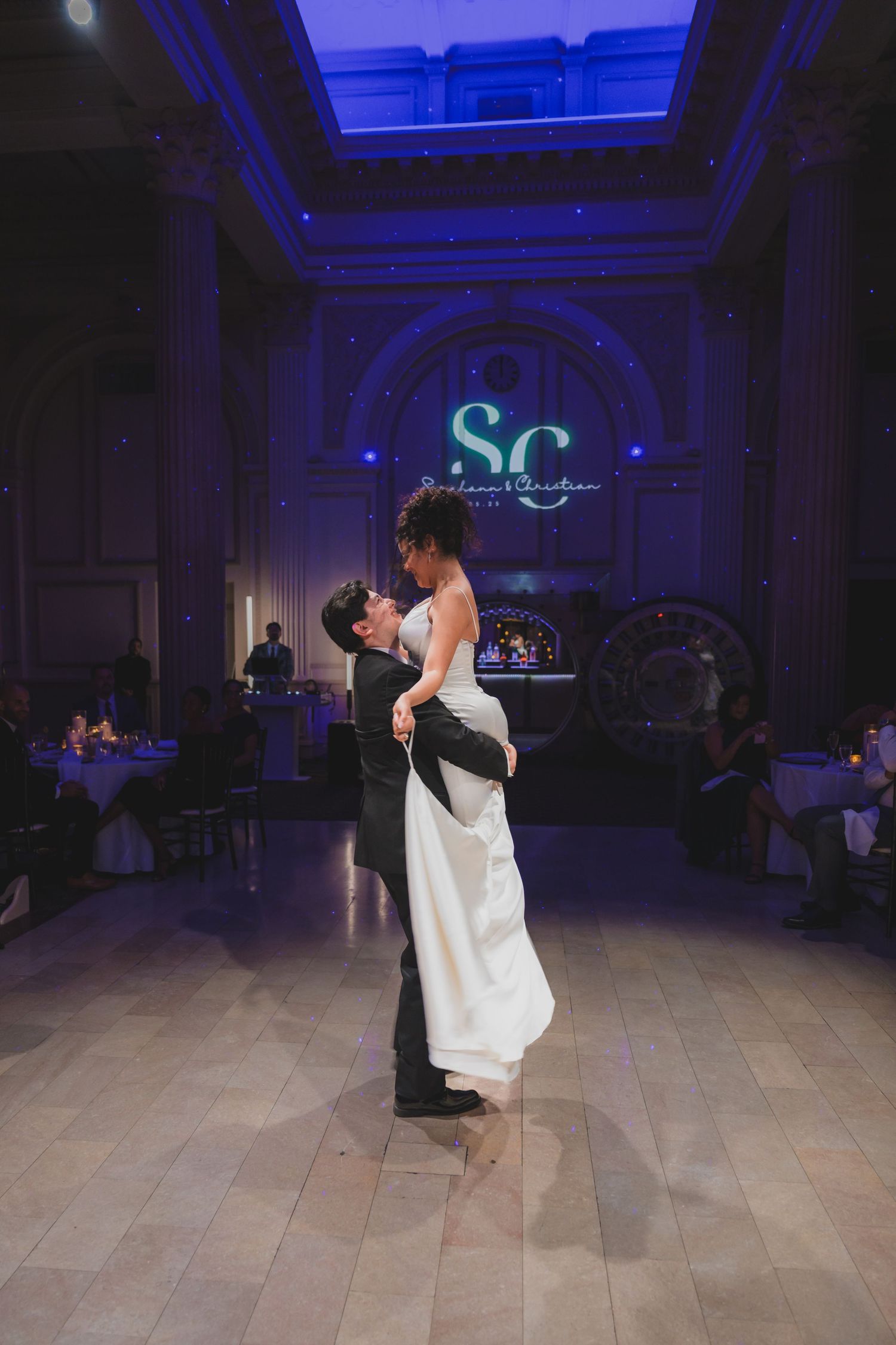 Groom lifts bride during first dance in grand ballroom with monogram SC light display.
