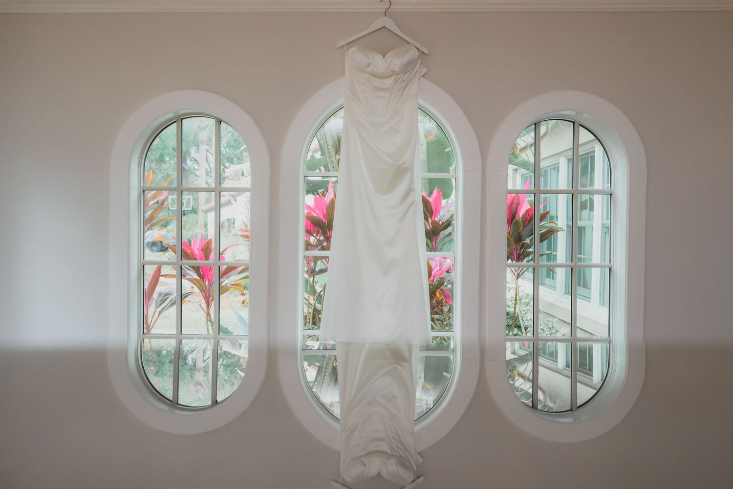 White wedding dress hanging on a hanger in front of three arched windows with tropical pink flowers outside.