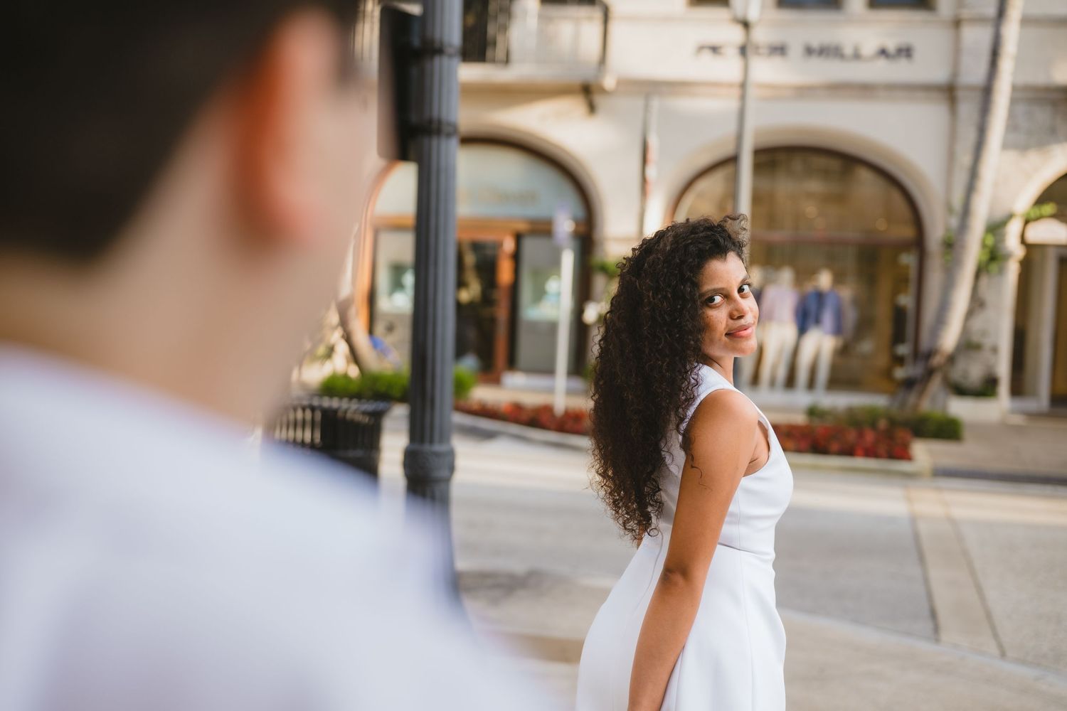 Woman in white dress glances back smiling at partner on sunny urban street.