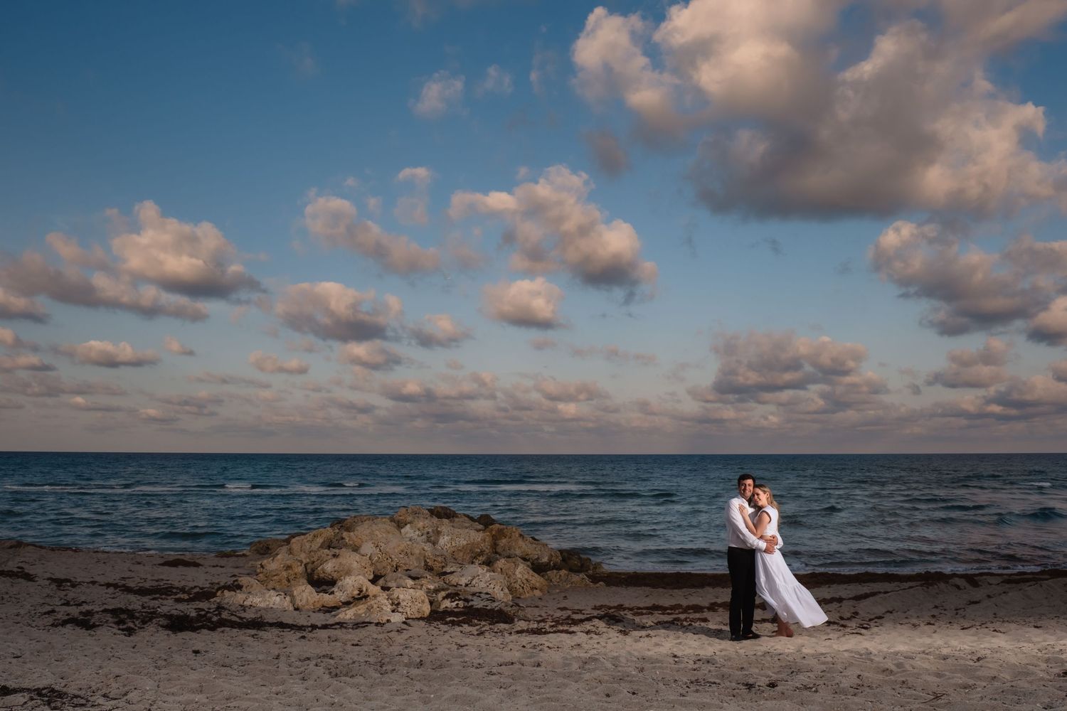 Couple embracing on rocky beach with dramatic cloudy sky and ocean backdrop.