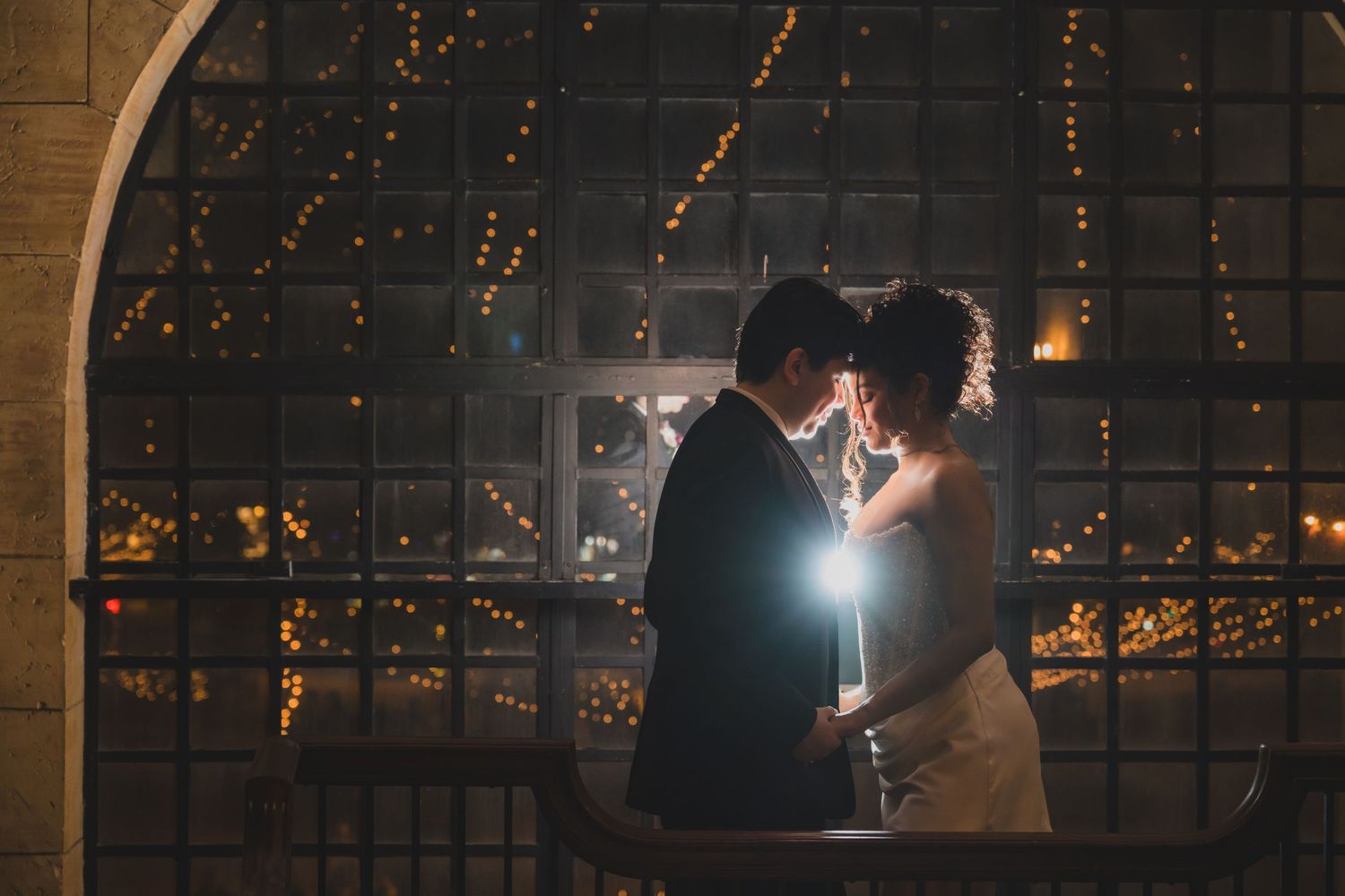 A couple shares a romantic moment in front of a glowing window with fairy lights at their wedding reception.