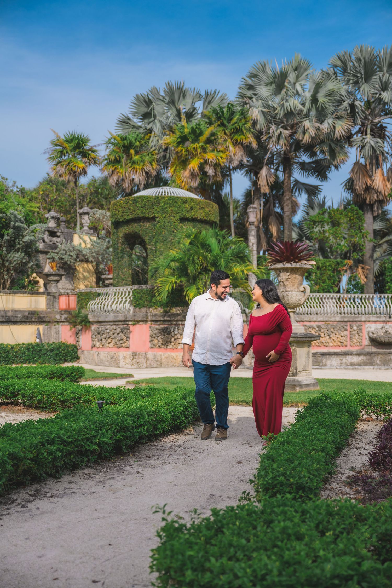Couple walking hand in hand through manicured garden paths with palm trees and ornate hedges.